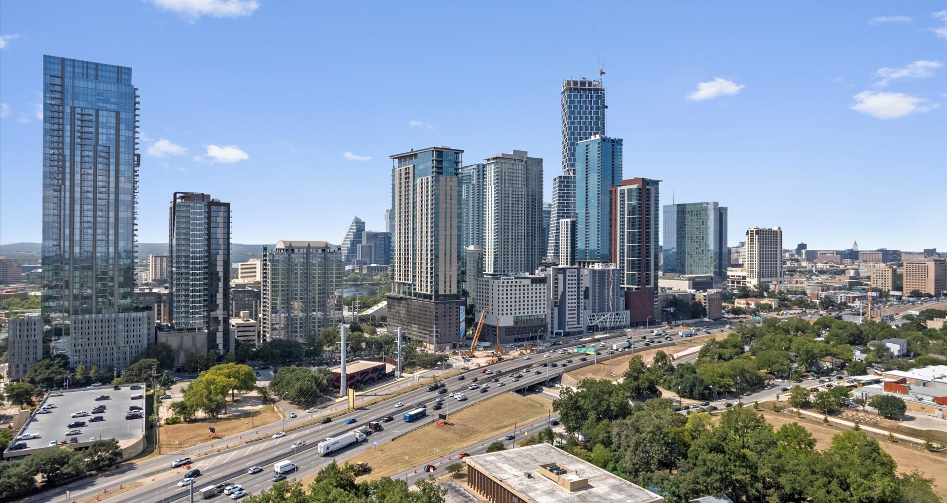 An aerial view of a bustling city skyline featuring numerous modern high-rise buildings under a clear blue sky, with a multi-lane highway winding through the foreground and lush green trees lining its edges.