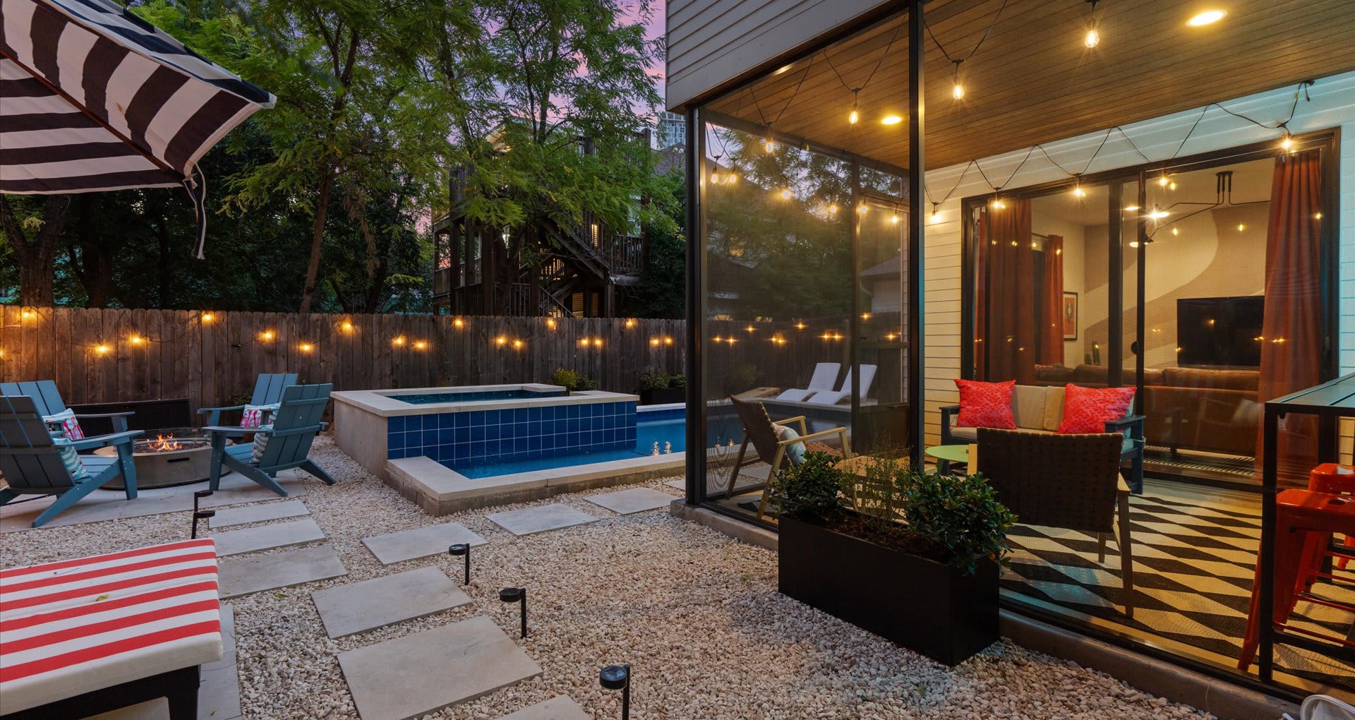 Expansive evening backyard view showcasing a lit pool, a fire pit area with chairs, a screened porch with glowing lights, and string lights draped across the wooden fence, creating a festive atmosphere.