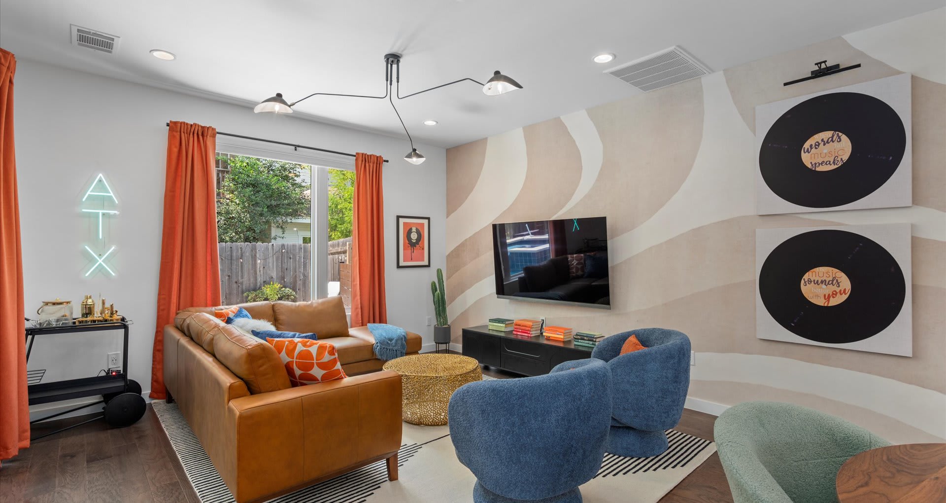 Stylish living room with a large wall-mounted TV, abstract wall mural, and two large vinyl record art pieces, complemented by a tan leather sectional and blue swivel chairs.