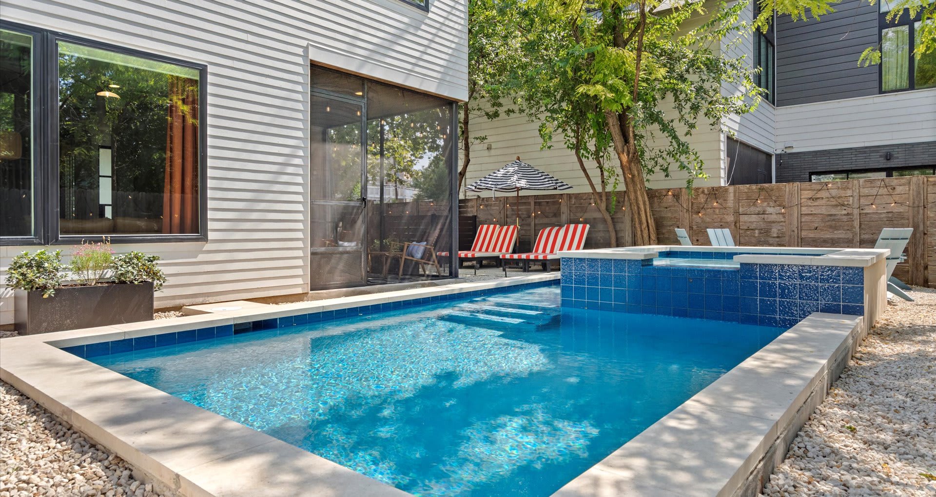 Modern rectangular pool with blue tiled interior and integrated hot tub, surrounded by light-colored paving and white gravel, with lounge chairs and an umbrella in the background.