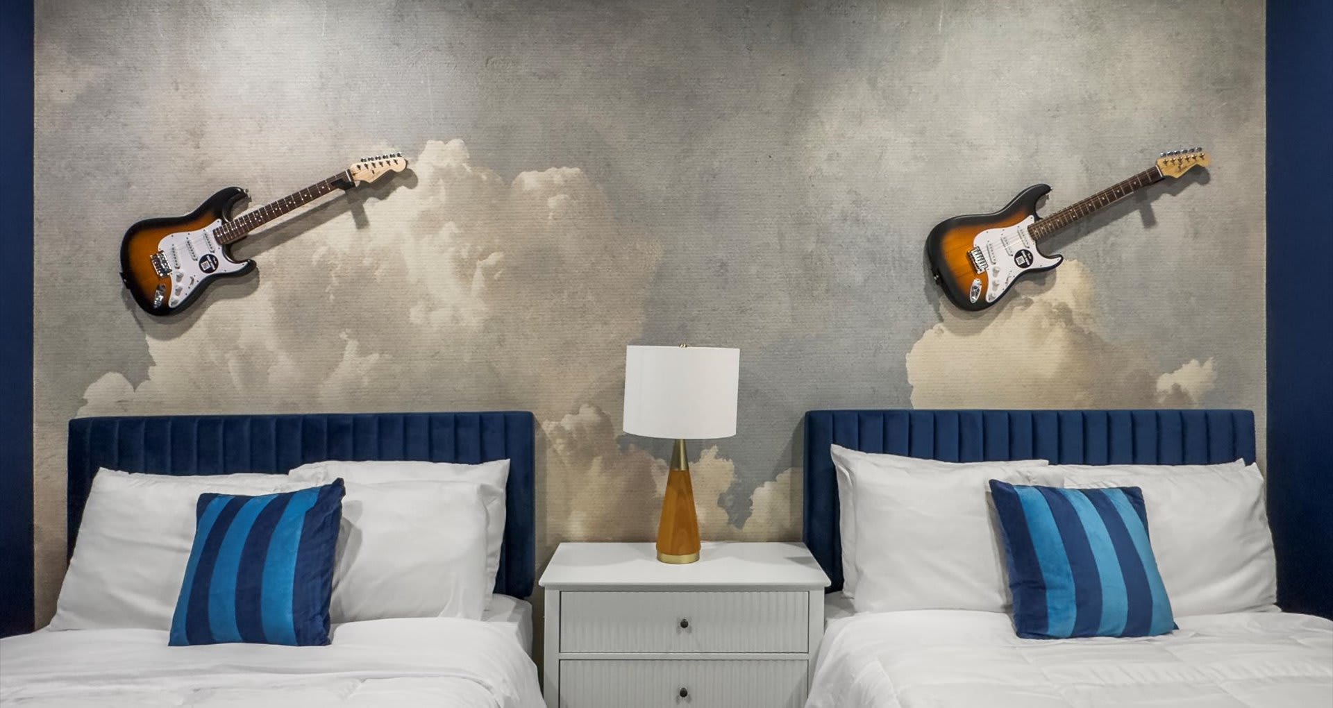 Close-up of two twin beds with blue velvet headboards and white bedding, adorned with blue striped pillows, separated by a white nightstand with a modern lamp. The gray cloud-patterned accent wall with guitar decor is prominent.