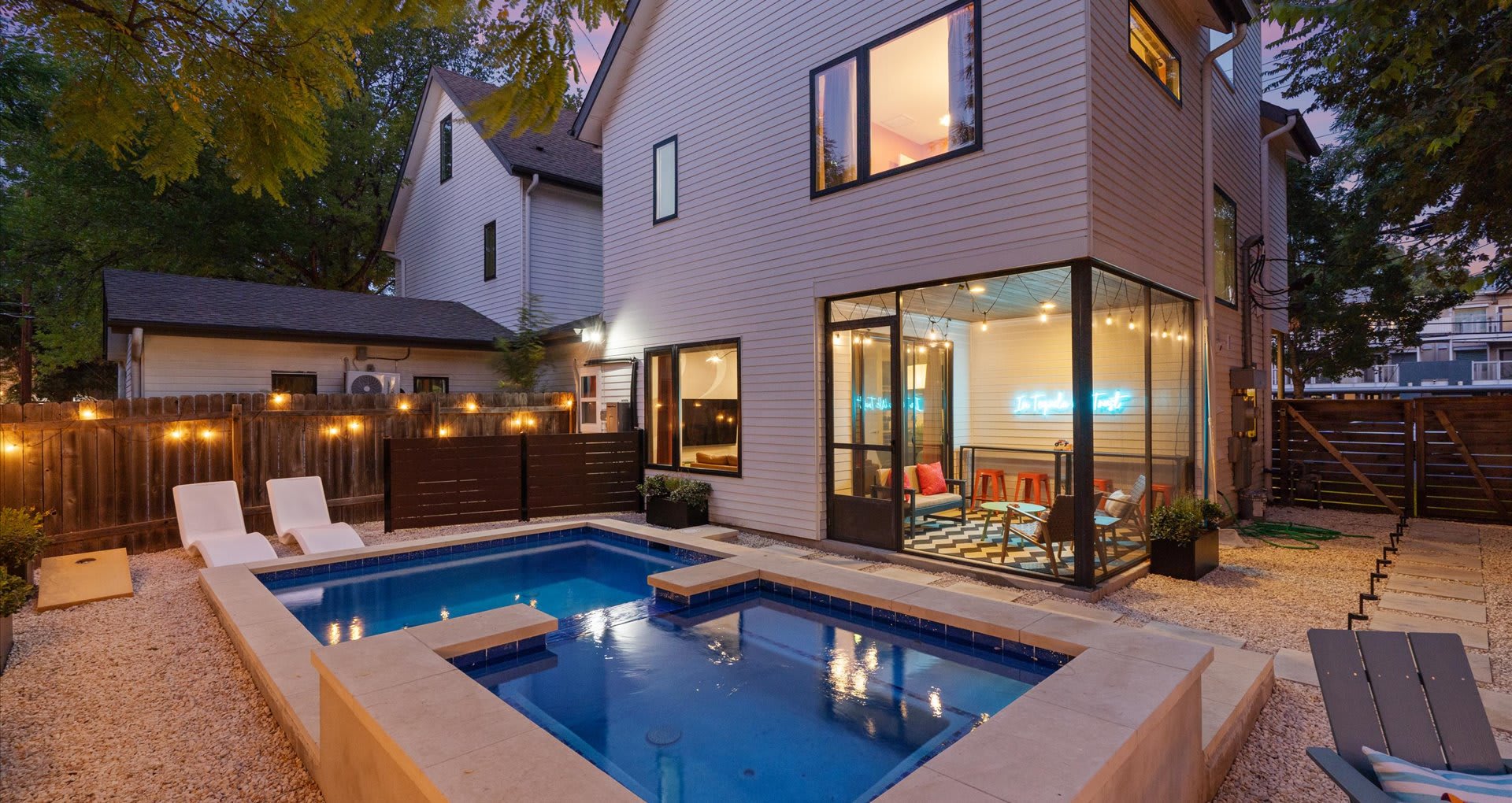 Evening view of the backyard featuring a lit pool and hot tub, a modern white house with a glowing screened porch, and string lights illuminating the wooden fence, creating a warm ambiance.