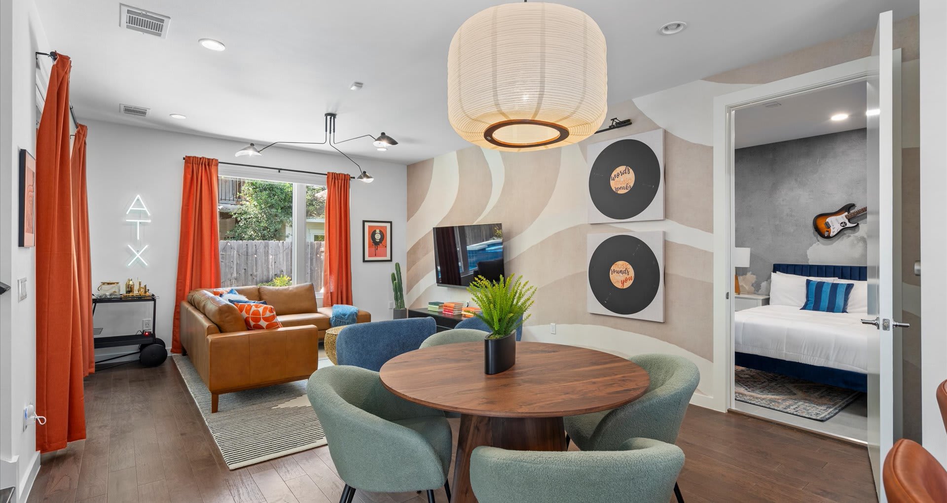 Dining area with a round wooden table and green upholstered chairs, illuminated by a large paper lantern, with the living room to the left and a bedroom entrance visible on the right.