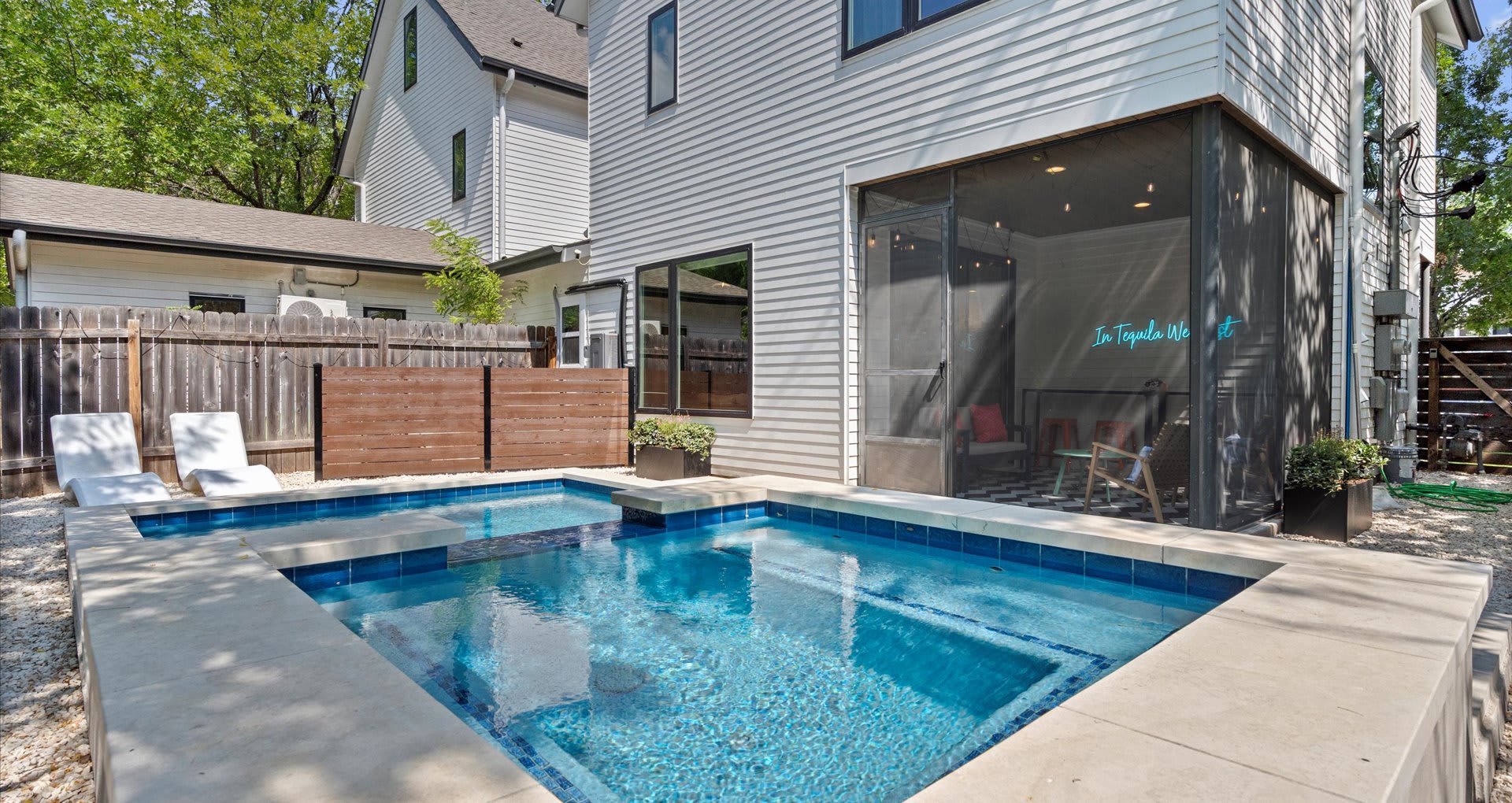 Sunny backyard featuring a rectangular pool with an integrated hot tub, surrounded by light gravel, a modern white house with a screened-in porch, and a wooden fence, perfect for relaxation.