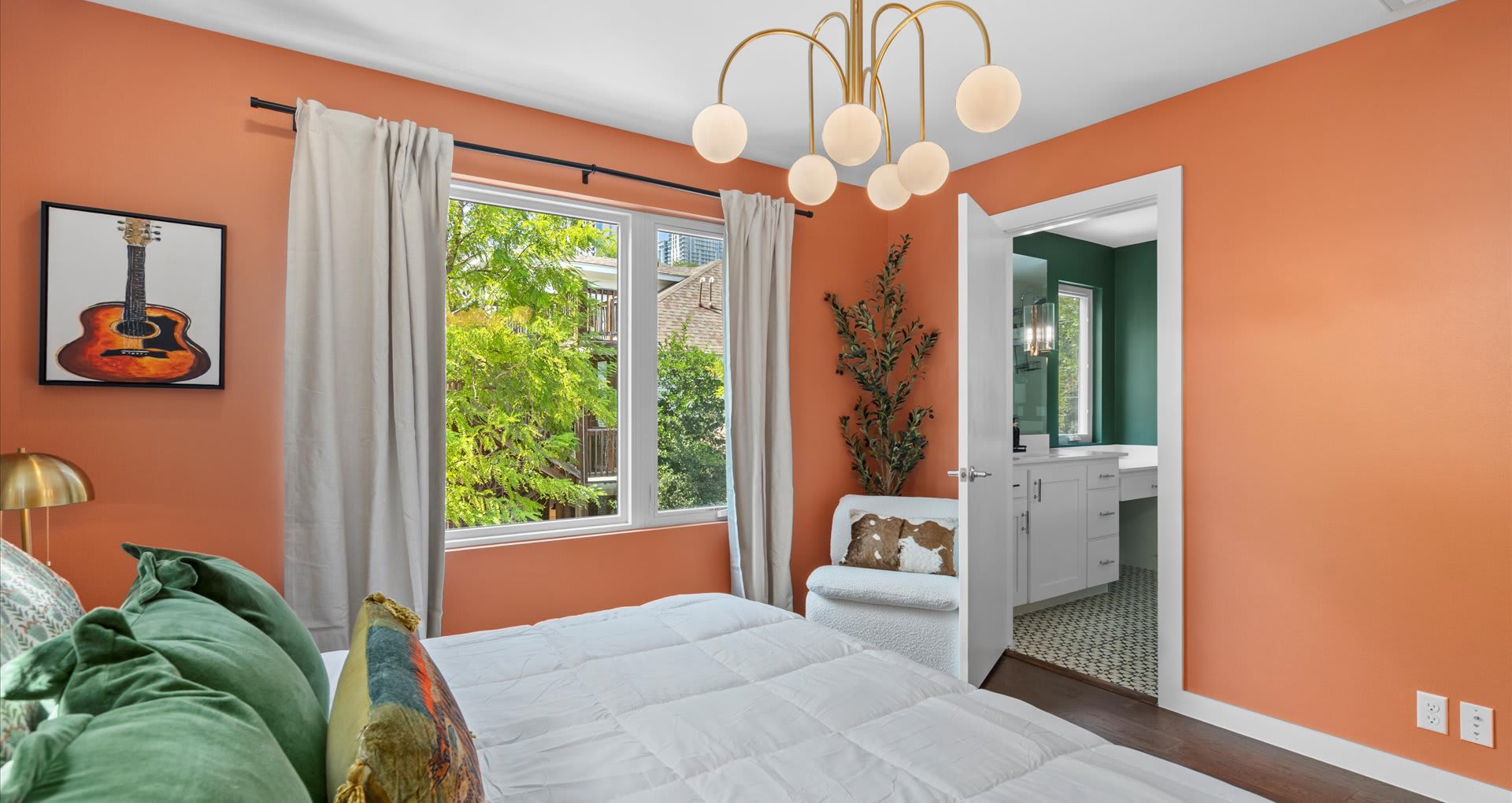 An expansive view of bedroom-3, featuring an orange accent wall, a modern brass chandelier, and a large window overlooking lush greenery. A white armchair and a doorway leading to a bathroom are also visible.