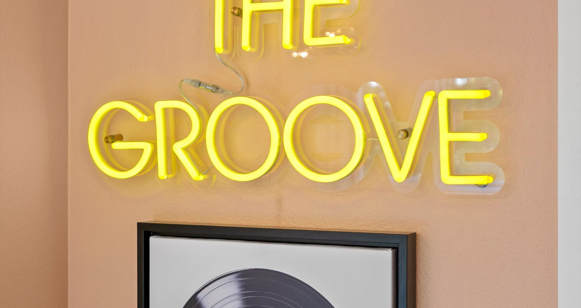 A close-up of a bright yellow neon sign on a peach wall, spelling out "FEEL THE GROOVE" above a framed record album, adding a retro touch to the bedroom decor.