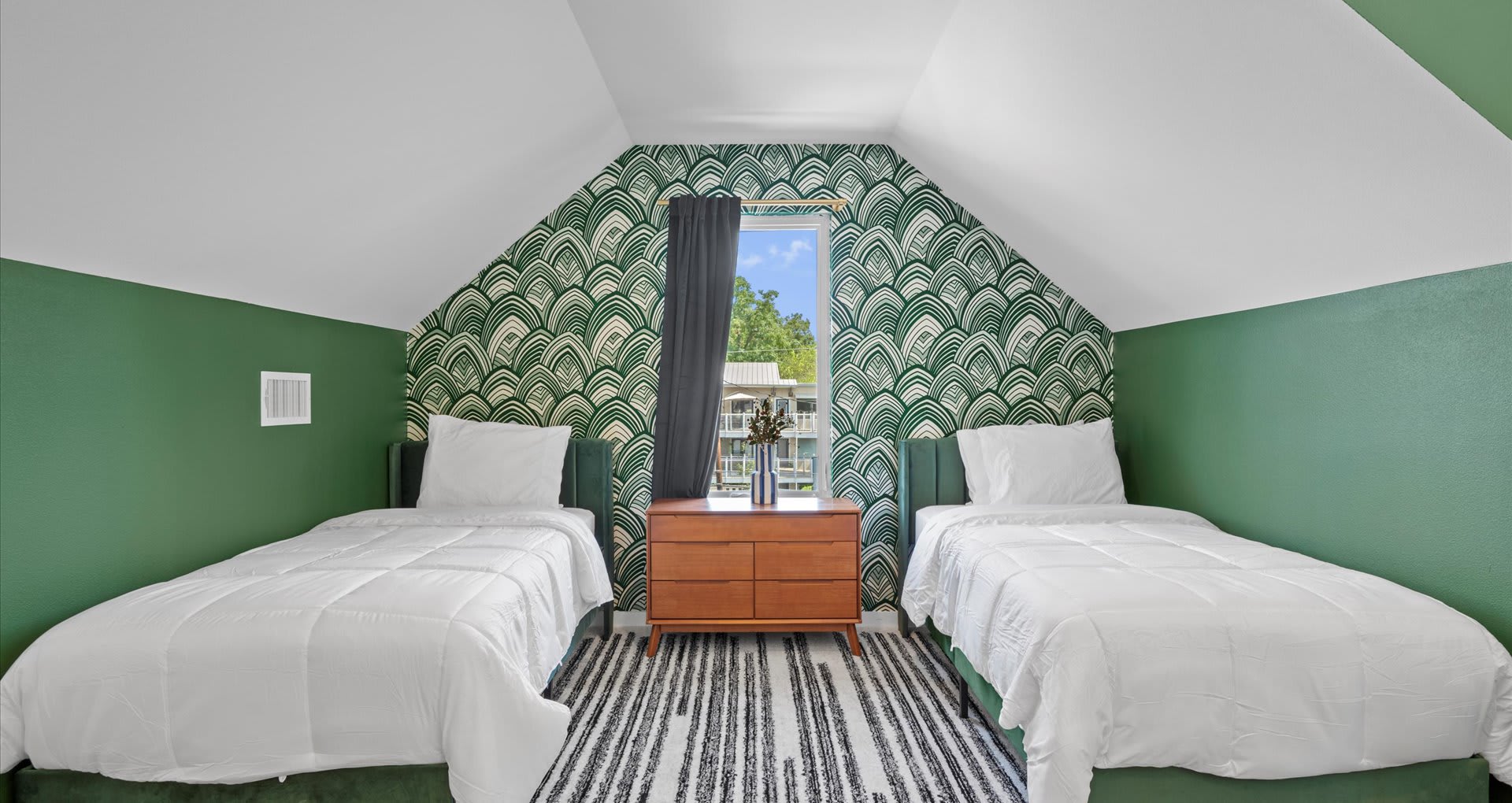 A cozy attic bedroom area with two twin beds, a wooden dresser between them, and a green patterned wallpaper accent wall, under a sloped white ceiling with a window.