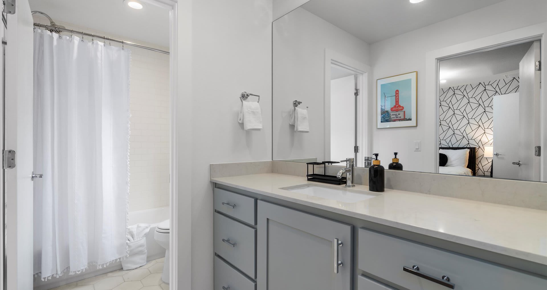 A modern bathroom featuring a light gray vanity with a white countertop and a large mirror. A shower/tub combination with white subway tiles and a white shower curtain is visible to the left.
