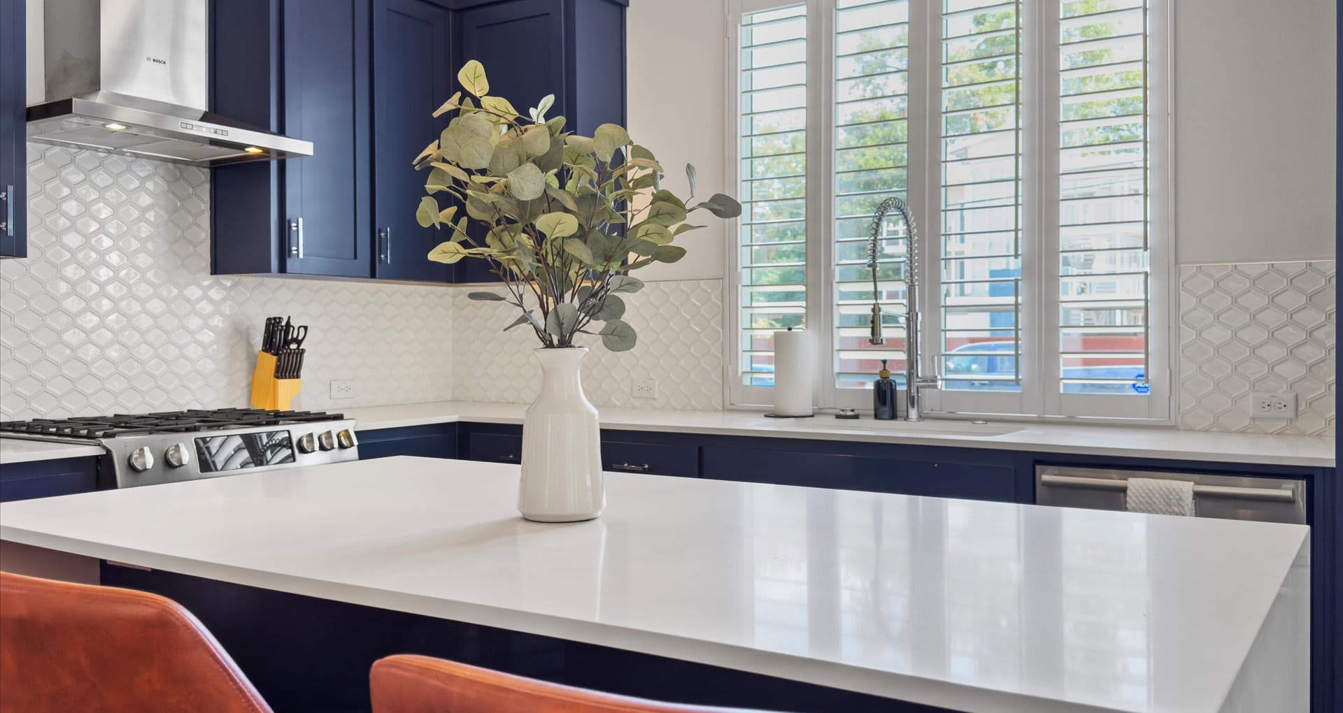 Modern kitchen island with a white quartz countertop and a decorative vase, featuring a white scalloped tile backsplash and a large window with white plantation shutters.