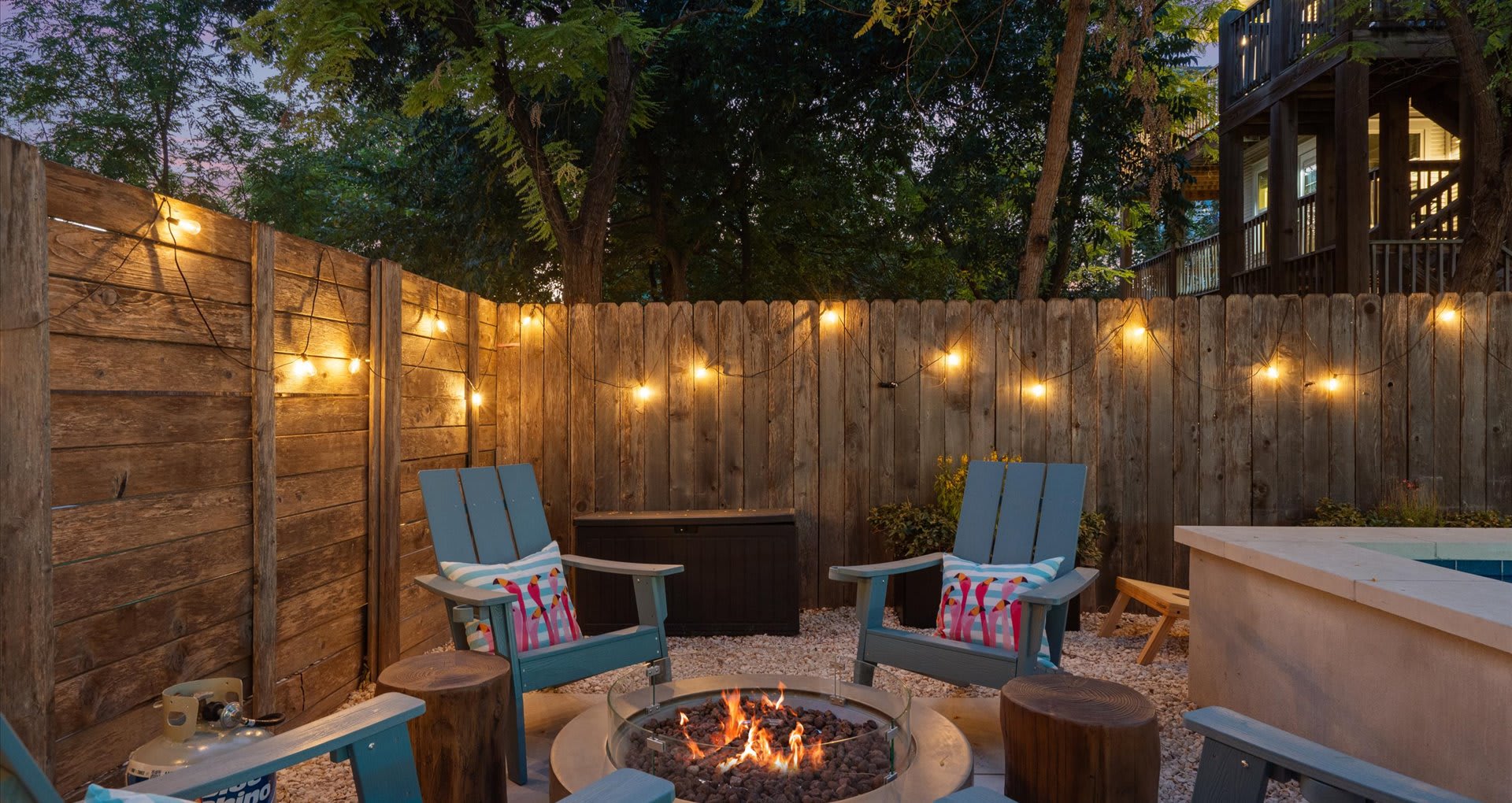 Cozy evening scene around a fire pit with dancing flames, surrounded by blue Adirondack chairs and wooden stumps, all illuminated by string lights along a rustic wooden fence.