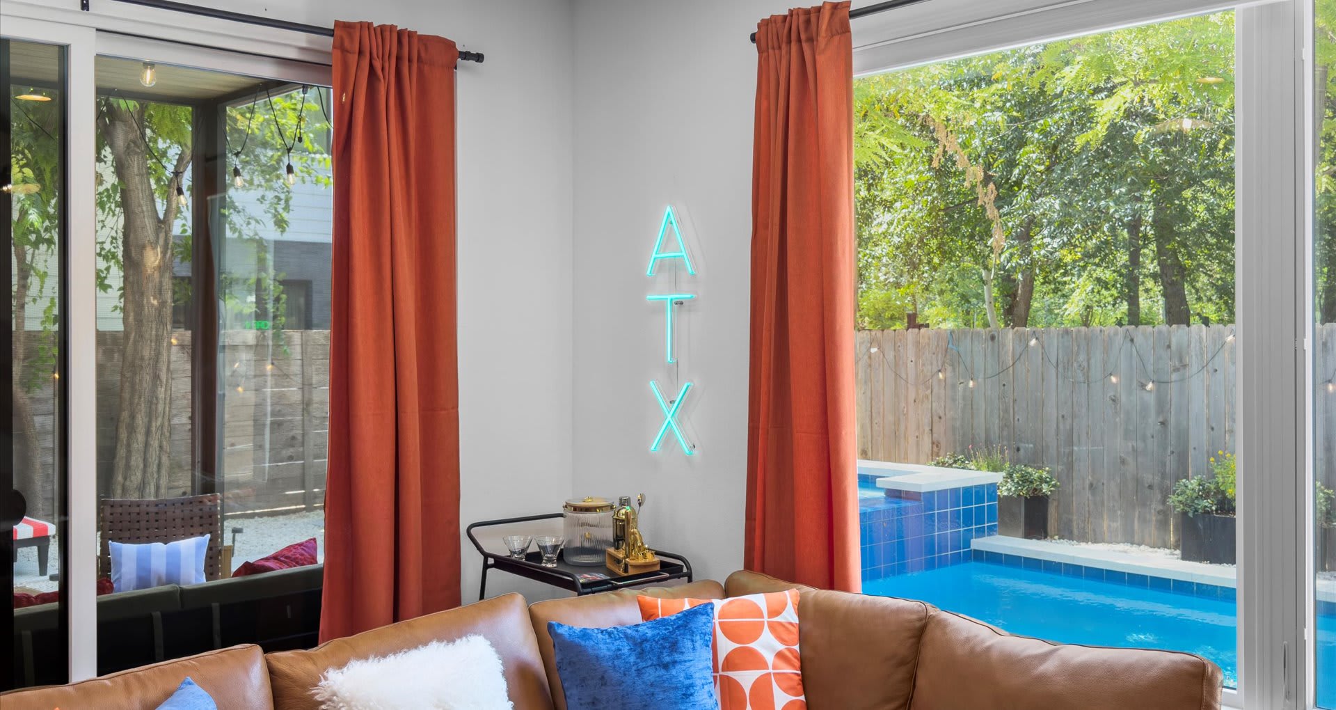 Cozy corner of the living room featuring a neon "ATX" sign above a bar cart, with large windows offering a direct view of the outdoor pool and backyard.