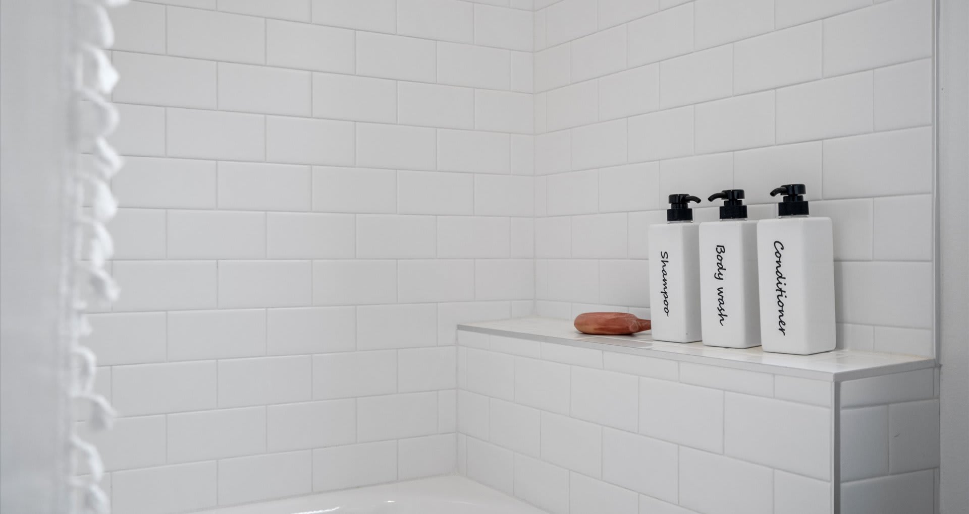 A close-up of a white subway-tiled shower with a built-in shelf. Three labeled bottles of shampoo, body wash, and conditioner, along with a bar of soap, are neatly arranged on the shelf.