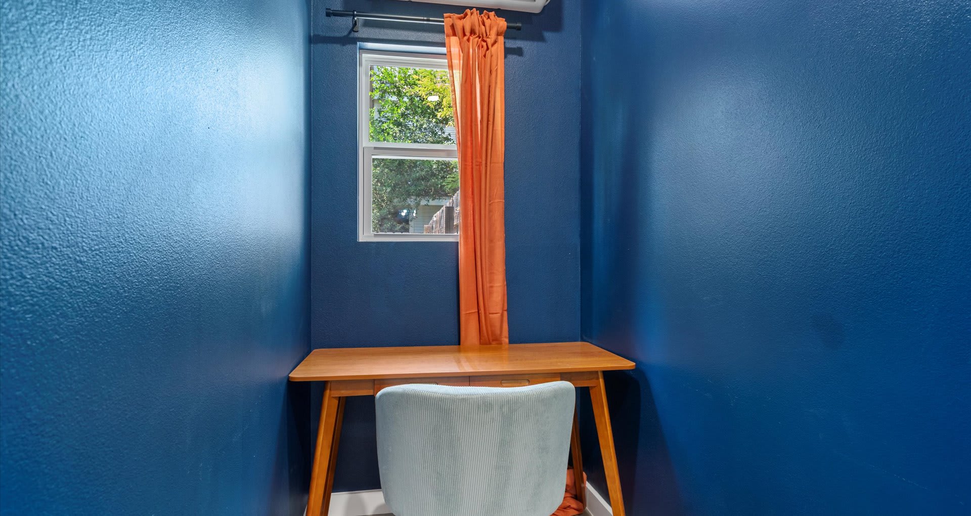 Cozy desk nook with bold blue walls, a light wood desk, and a light blue upholstered chair, illuminated by natural light from a small window with orange curtains. A mini-split AC unit is mounted above the window.