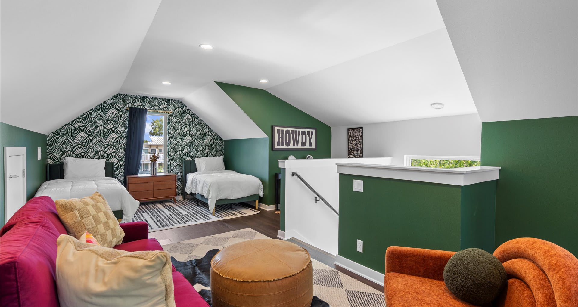 An attic bedroom and lounge area with a sloped ceiling, featuring two twin beds against a green patterned wallpaper accent wall, a red sofa, and a 'HOWDY' sign above a half-wall.