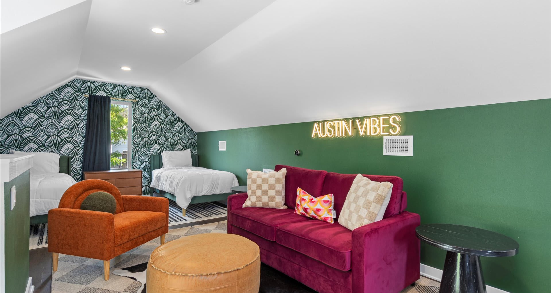 A vibrant attic bedroom and lounge area with a sloped ceiling, featuring a red sofa, an orange armchair, and a neon 'AUSTIN VIBES' sign on a dark green wall.