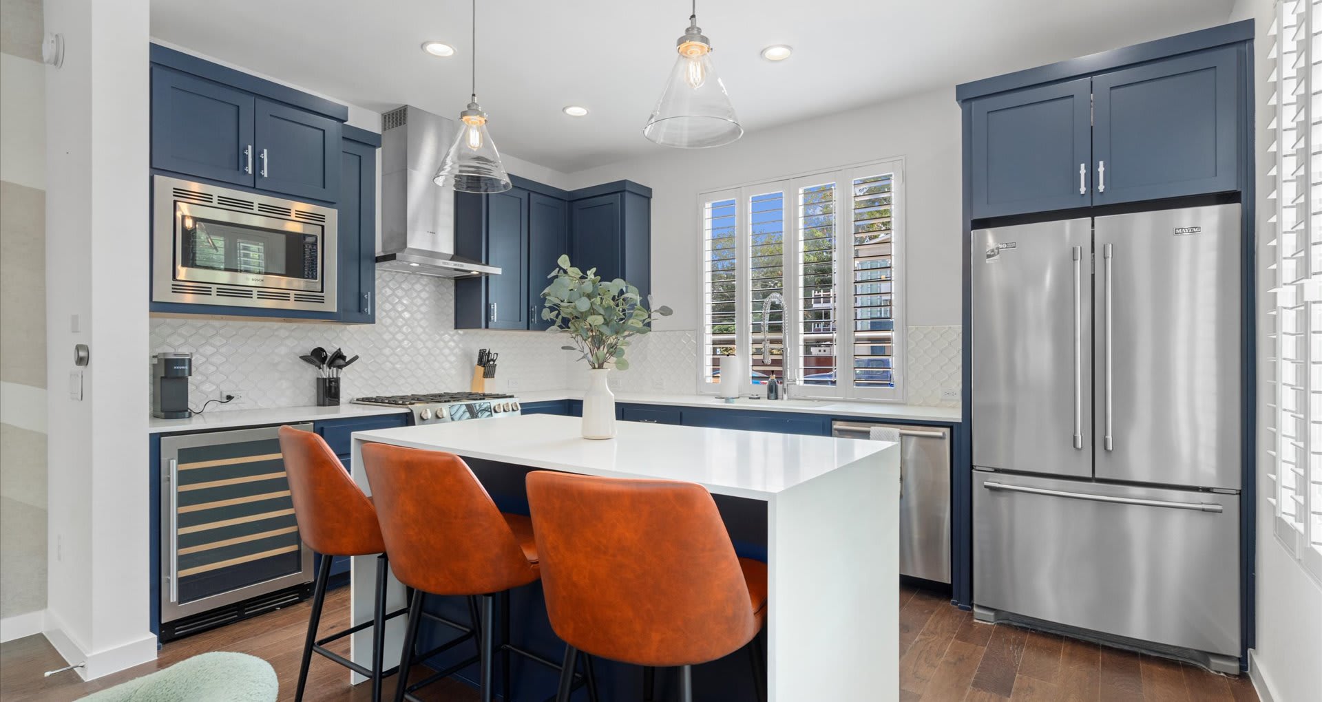 Contemporary kitchen with dark blue shaker cabinets, white quartz countertops, stainless steel appliances, and a central island with three orange bar stools.