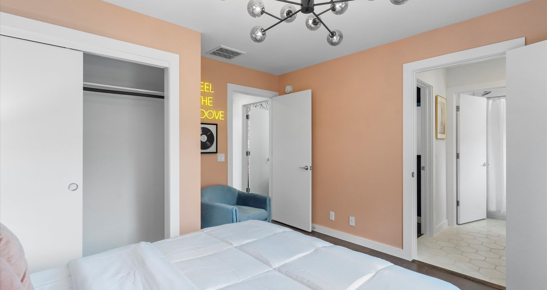 An overview of bedroom-1, showcasing peach walls, a white bed, and a vibrant yellow neon sign on the wall. A light blue armchair sits in the corner, and a modern multi-bulb chandelier hangs from the ceiling.