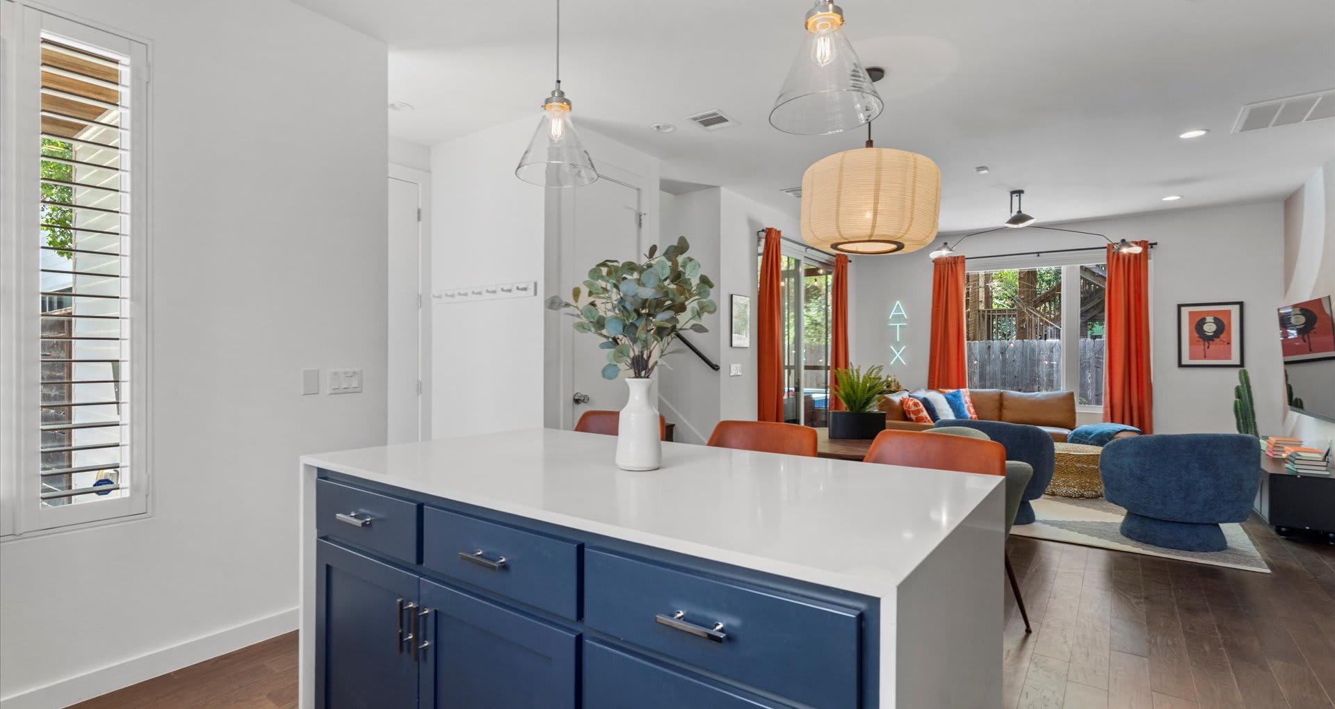 Kitchen island with dark blue cabinetry and a white countertop, featuring a decorative vase with greenery. The open-concept living area with orange curtains, modern lighting, and a neon 'ATX' sign is visible in the background.