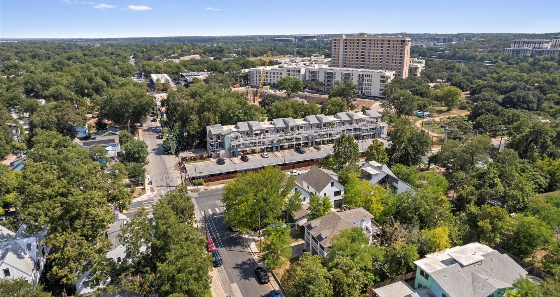An aerial view showcasing the property and its surrounding neighborhood, characterized by lush green trees, residential buildings, and a distant city skyline under a clear blue sky.