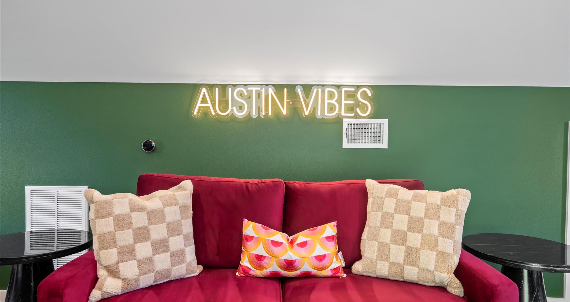 A close-up of a red velvet sofa adorned with patterned pillows, set against a dark green wall with a bright neon 'AUSTIN VIBES' sign above it, adding a playful touch.