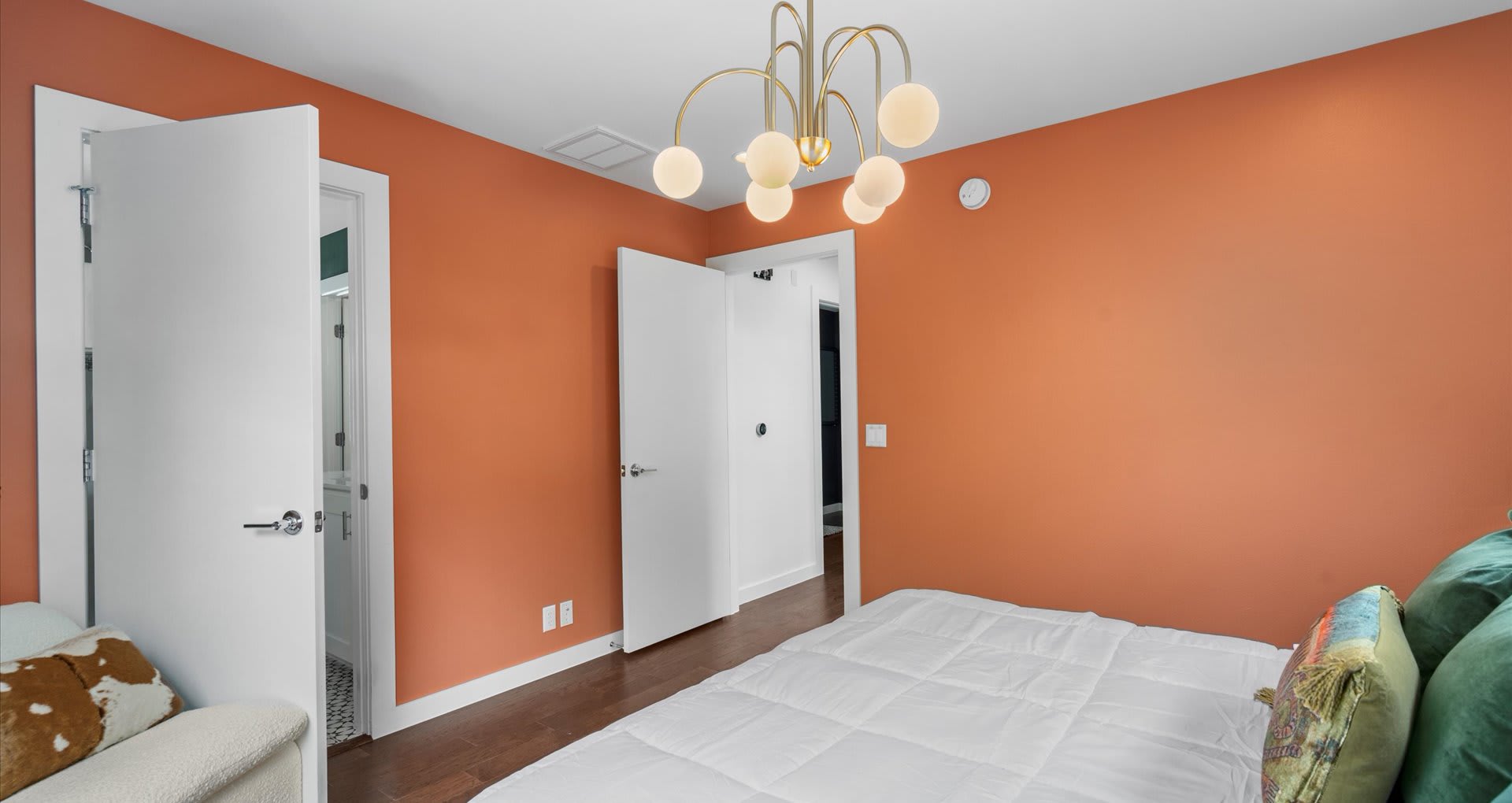 A vibrant bedroom featuring an orange accent wall, dark wood floors, and a white bed with colorful pillows, illuminated by a modern gold chandelier with globe lights.