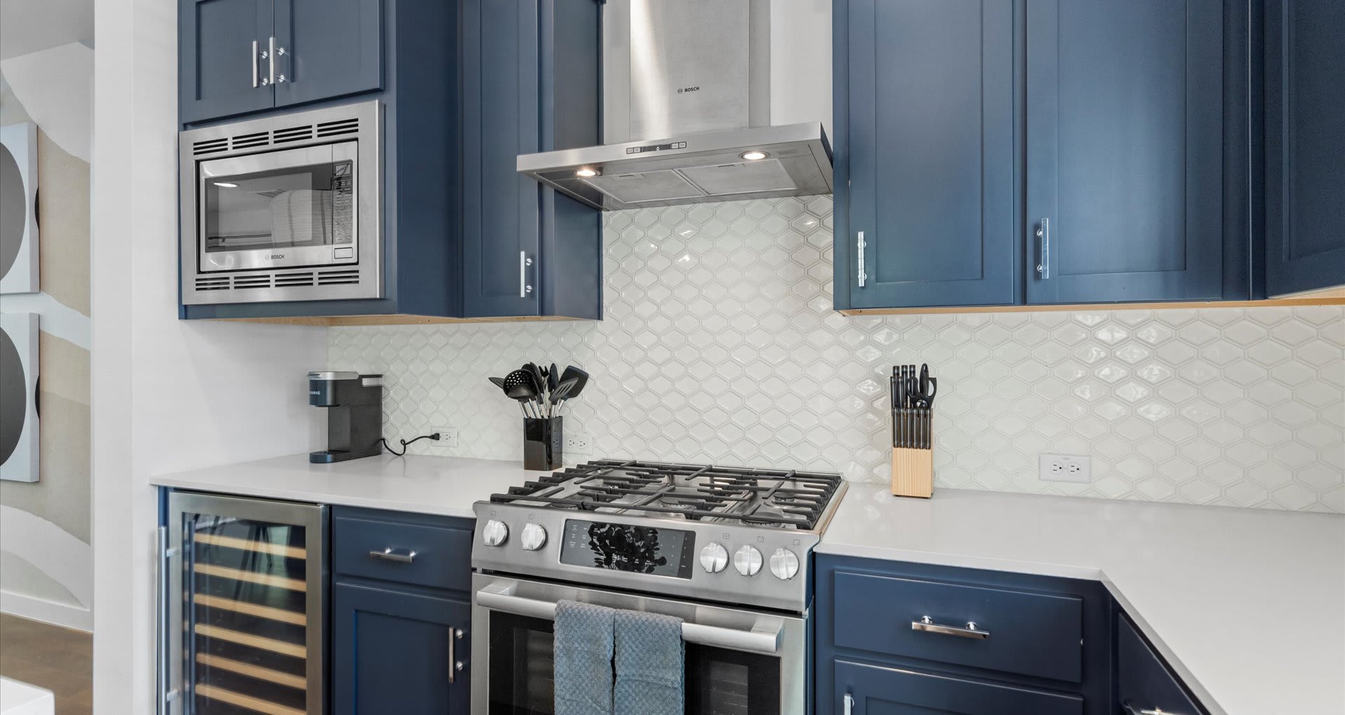 Modern kitchen featuring dark blue shaker-style cabinets, white quartz countertops, a white geometric tile backsplash, and stainless steel appliances including a gas range, microwave, and range hood. A built-in wine cooler is visible on the left.