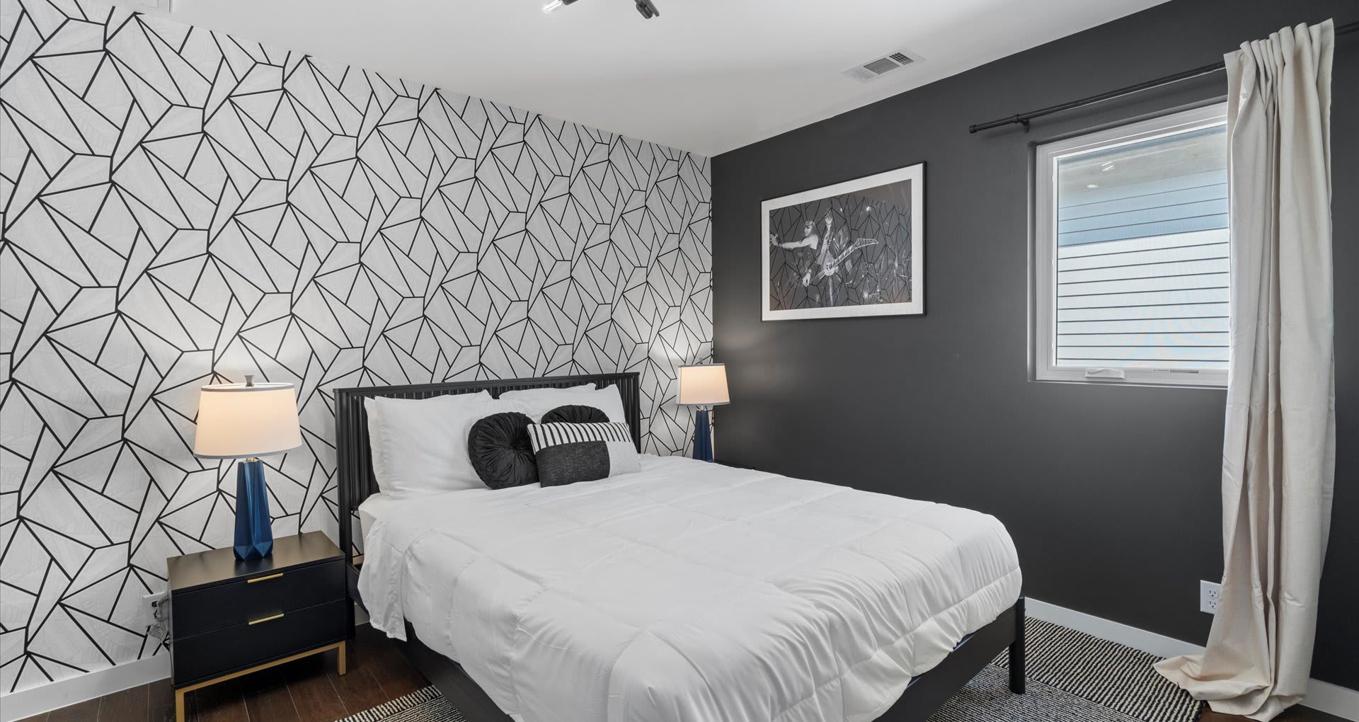 A bedroom with a striking black and white geometric patterned accent wall behind the bed, complemented by dark gray walls. Two black nightstands with blue-based lamps flank the bed.