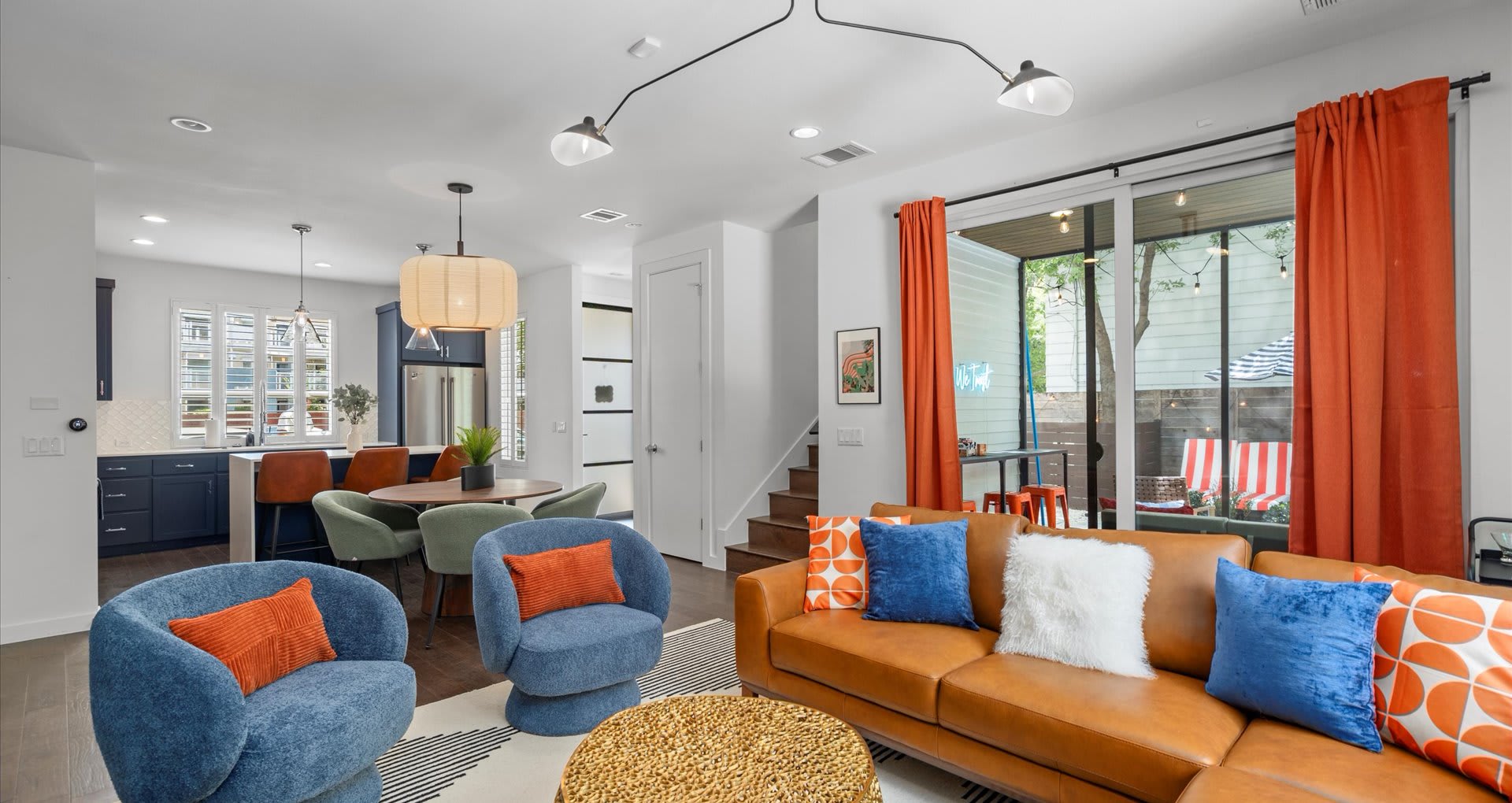 Bright open-concept living area with a tan leather sectional, blue accent chairs, a round wooden dining table with green chairs, and a glimpse of a modern blue kitchen.