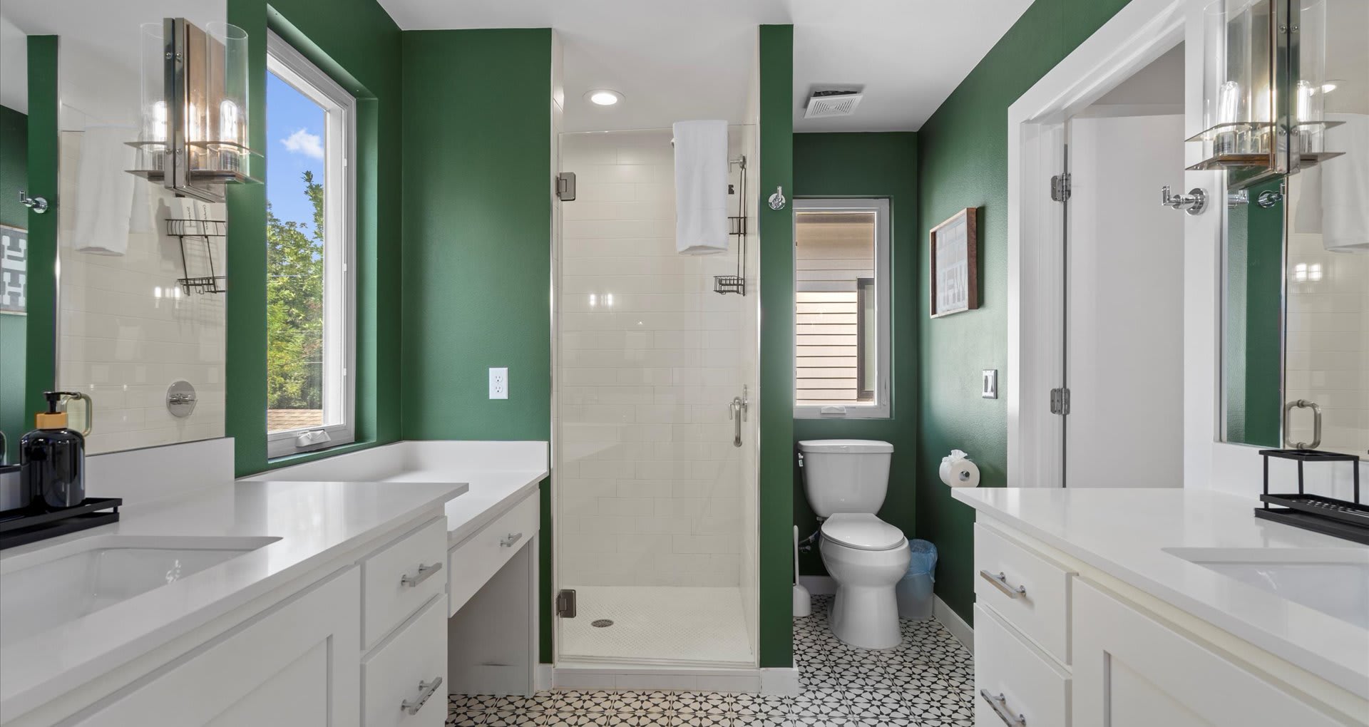 A spacious bathroom with dark green walls, a white toilet, a glass-enclosed shower with white subway tiles, and a white vanity, featuring black and white patterned floor tiles.