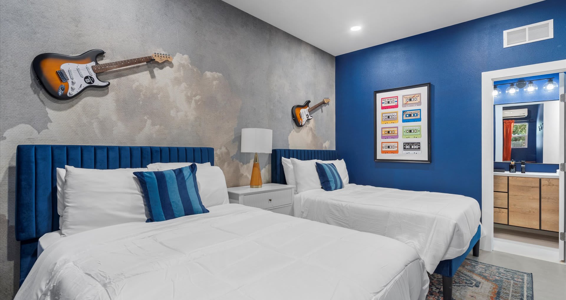 Spacious bedroom with two twin beds, blue velvet headboards, and white bedding, featuring a gray cloud-patterned accent wall with guitar decor and a bold blue wall with framed cassette tape artwork. An en-suite bathroom entrance is visible on the right.