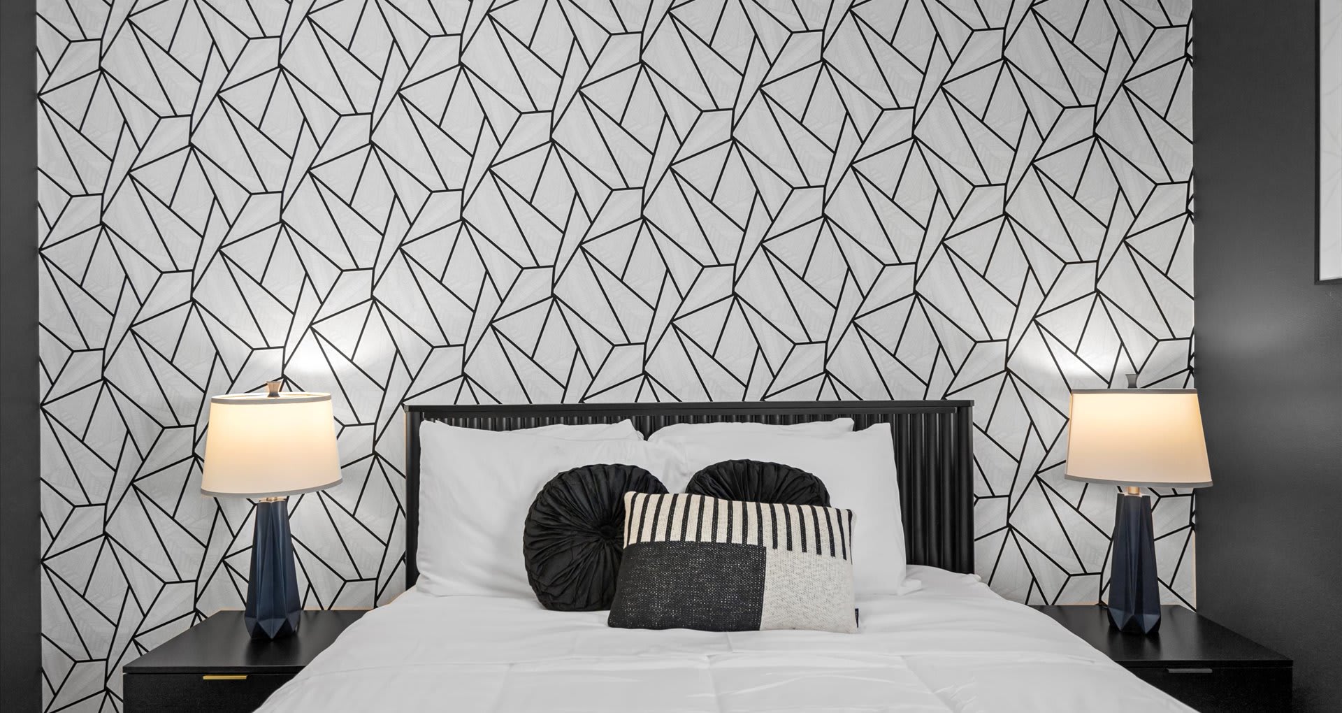 A close-up of the bed in bedroom-2, featuring a black headboard, crisp white bedding, and decorative black and white pillows. The geometric patterned accent wall provides a bold backdrop.