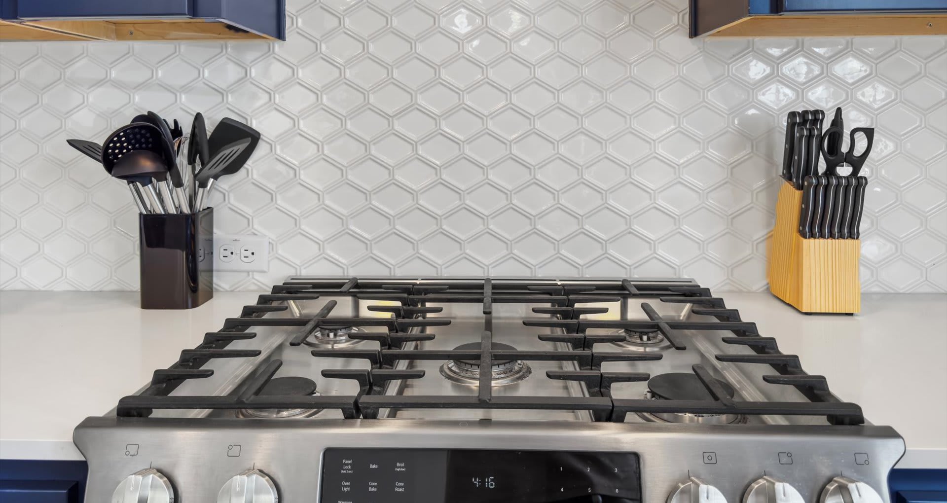 Close-up view of a stainless steel gas range with black grates and control knobs, set against a white geometric tile backsplash and white quartz countertop. A black utensil holder and knife block are on the counter.