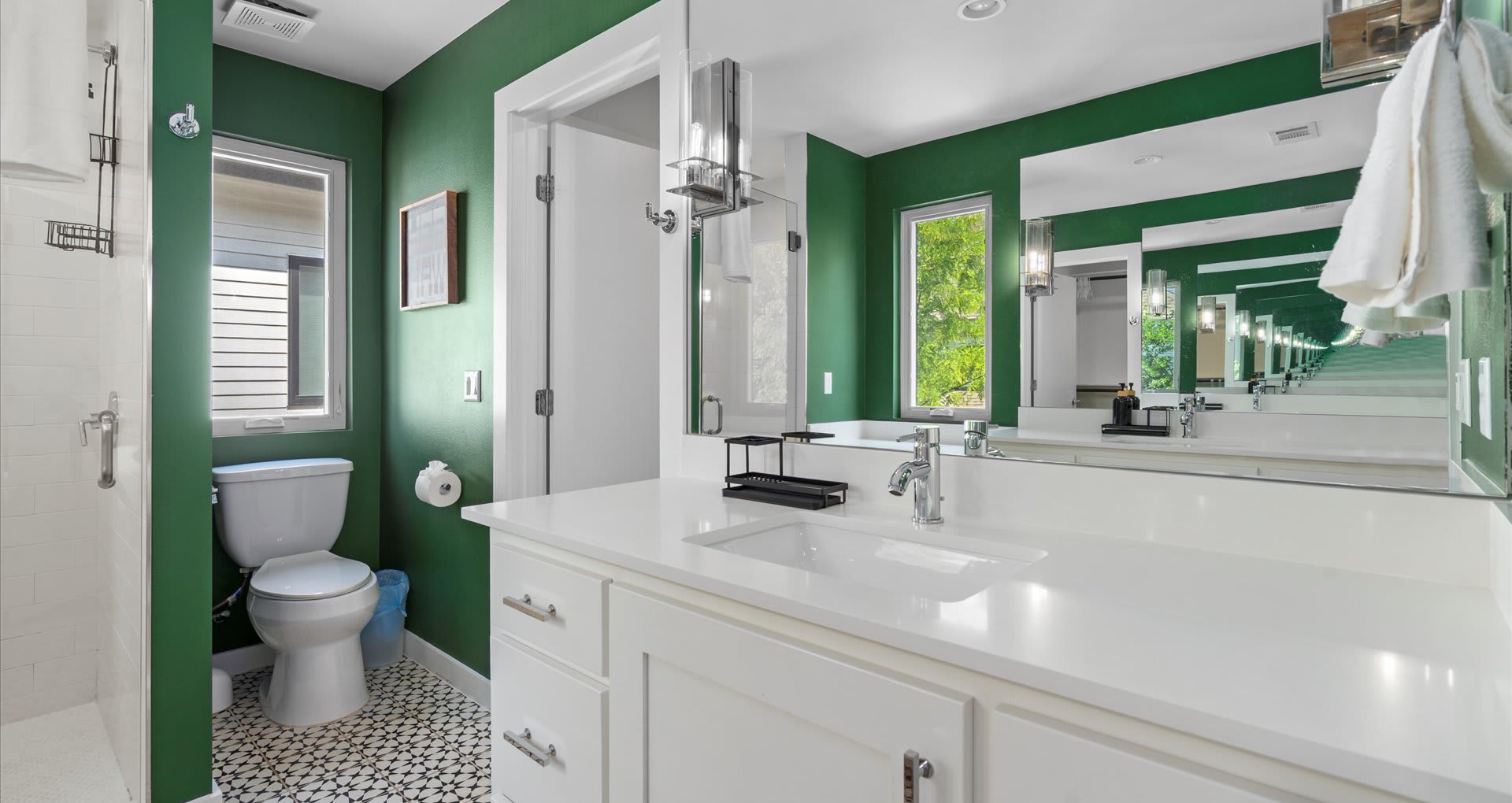 A bright bathroom with dark green walls, a white vanity with a single sink, a large mirror reflecting the room, and black and white patterned floor tiles, with a window providing natural light.