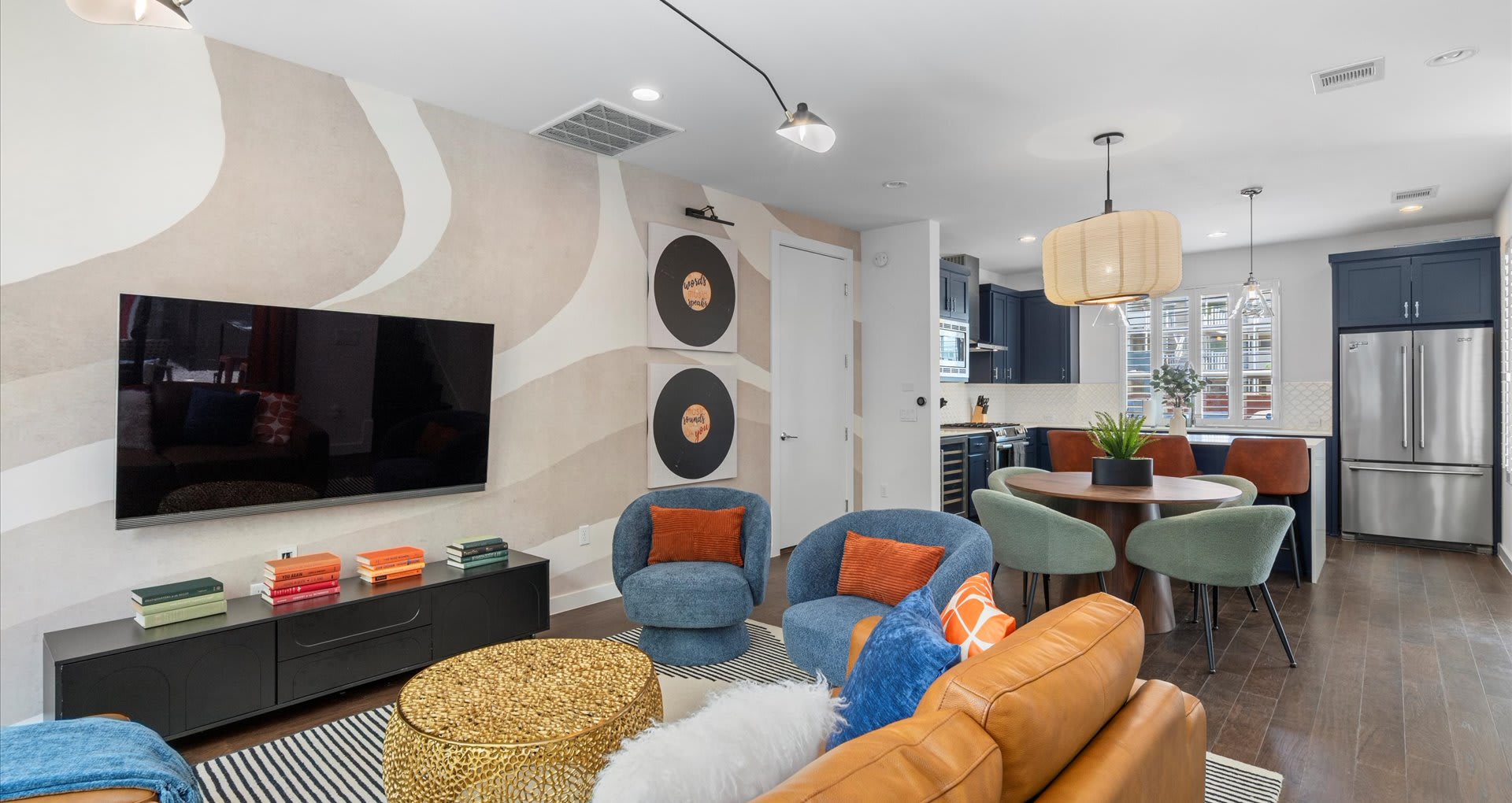 Vibrant living room with a tan leather sofa, blue accent chairs, and a woven coffee table, flowing into a dining area with a round table and a modern kitchen with blue cabinetry.