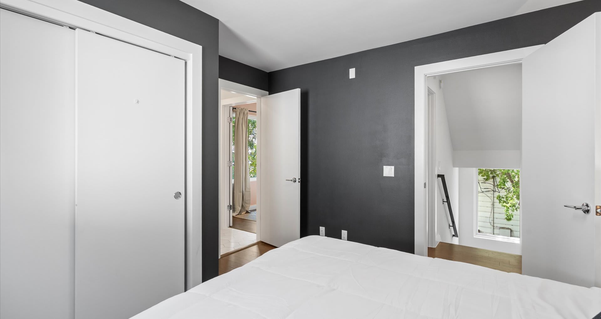 An alternate view of bedroom-2, showcasing dark gray walls, a white bed, and white sliding closet doors. A doorway leads to another area, possibly a hallway or balcony, with natural light.