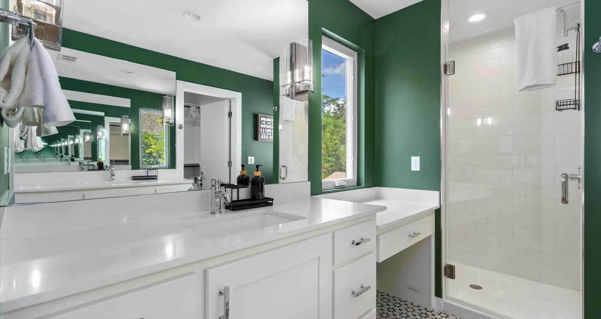 A modern bathroom with dark green walls, a long white double vanity with two sinks, a large mirror, and a glass-enclosed shower with white subway tiles and black and white patterned floor tiles.