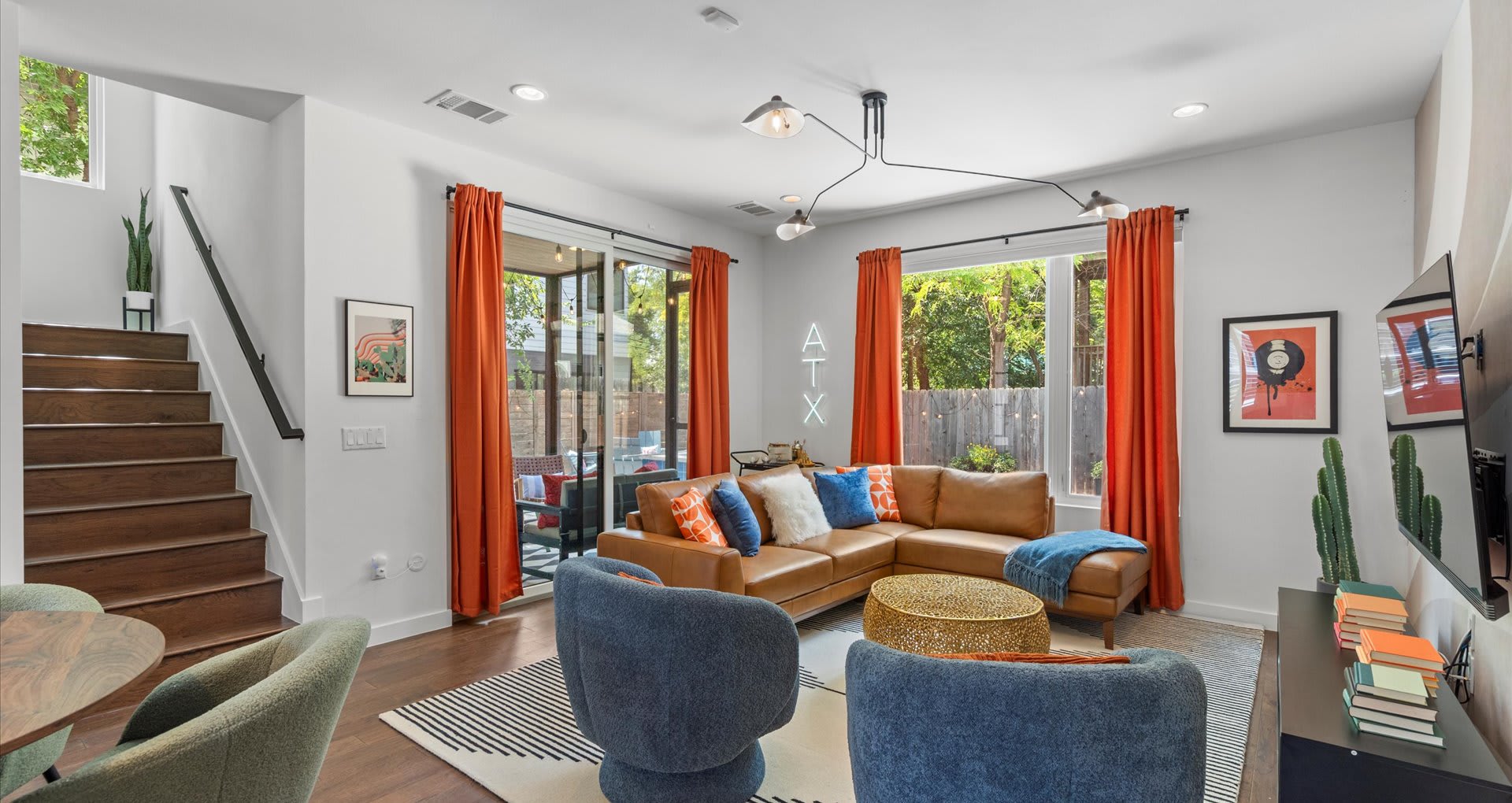 Vibrant living room featuring a large brown sectional sofa, two blue swivel chairs, a modern chandelier, and a wooden staircase, accented by orange curtains and an 'ATX' neon sign.