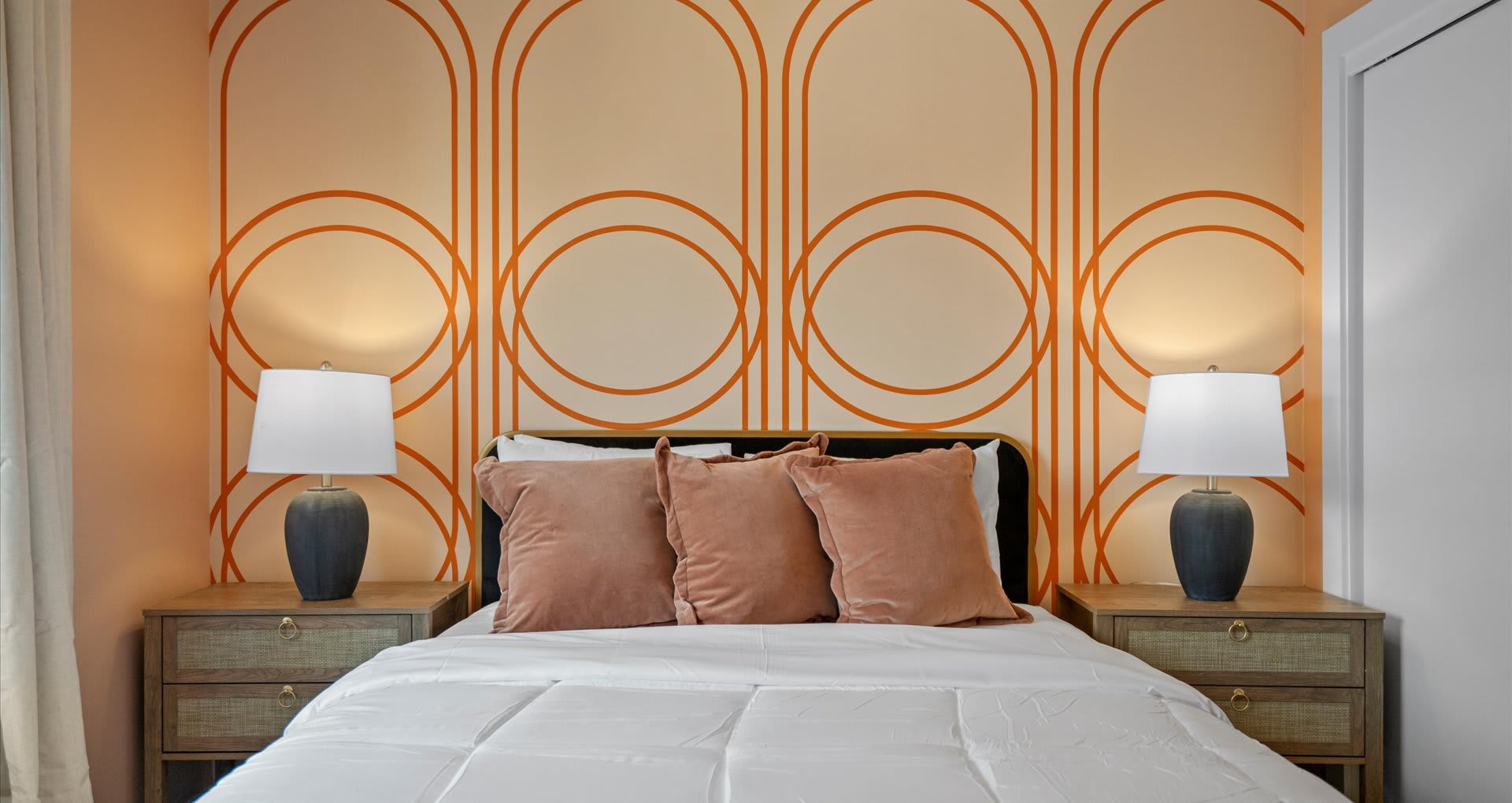 A bedroom featuring a bed with a black headboard, two nightstands with white lamps, and a peach-colored accent wall adorned with orange geometric patterns. Three pink velvet pillows add a pop of color.