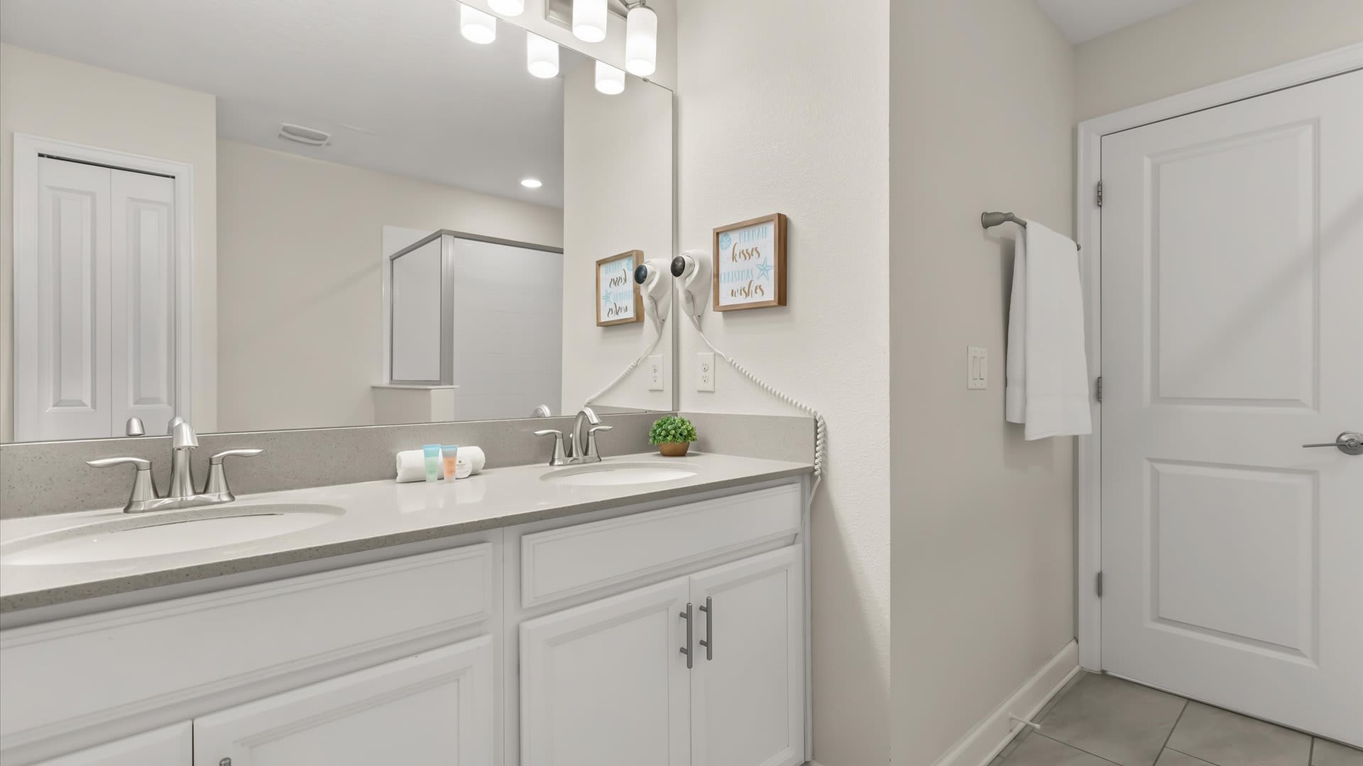 A bright bathroom features a double vanity with a light gray countertop, two sinks, and modern chrome faucets, illuminated by a strip light fixture above a large mirror. Two framed pictures and a hairdryer are mounted on the wall next to the vanity.