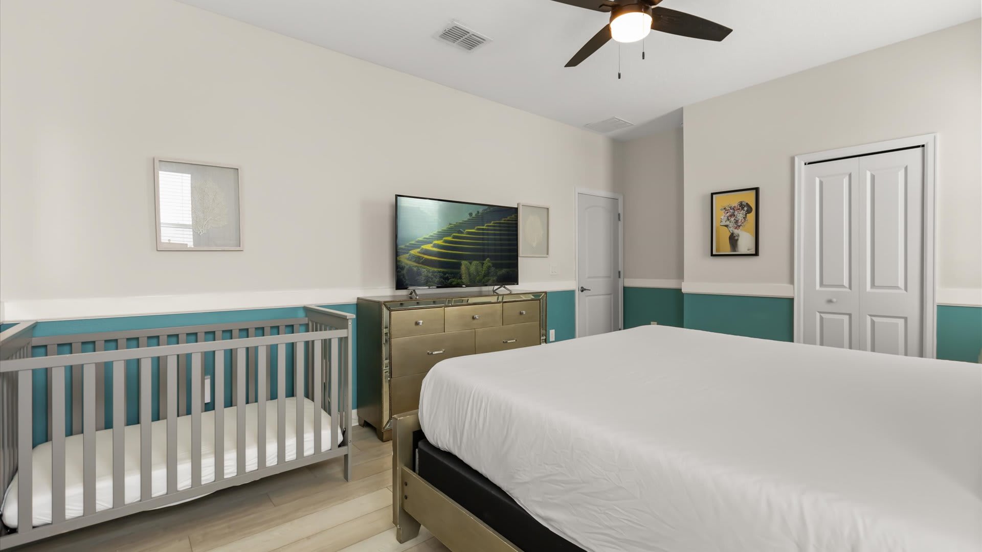 Another view of the bedroom, showcasing a large flat-screen TV atop a mirrored dresser, positioned next to the baby crib. The room maintains its two-tone wall design and light wood-look flooring.