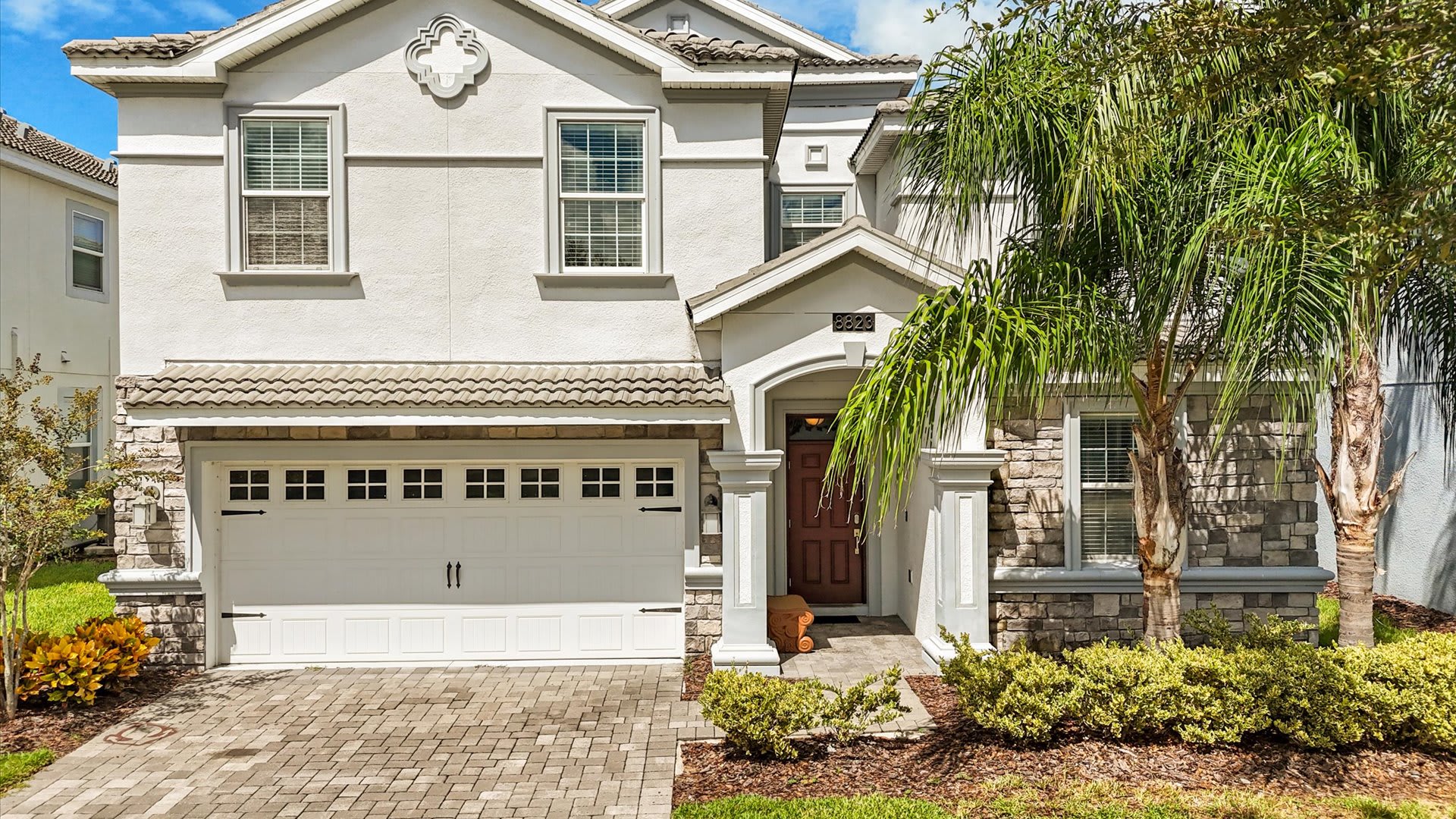 The front exterior of a two-story house features light gray stucco and stone facade, a two-car garage with a white door, and a covered entryway with columns. Lush green landscaping and a palm tree are visible.