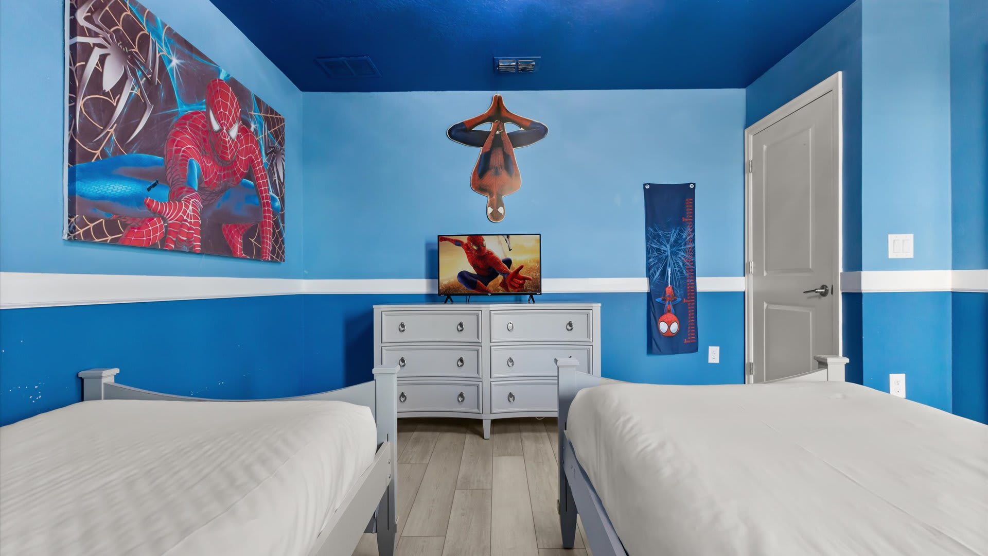 This Spiderman-themed bedroom showcases a white dresser with silver pulls, topped with a flat-screen TV displaying Spiderman, and an upside-down Spiderman decal on the blue wall above. Two twin beds are visible in the foreground.