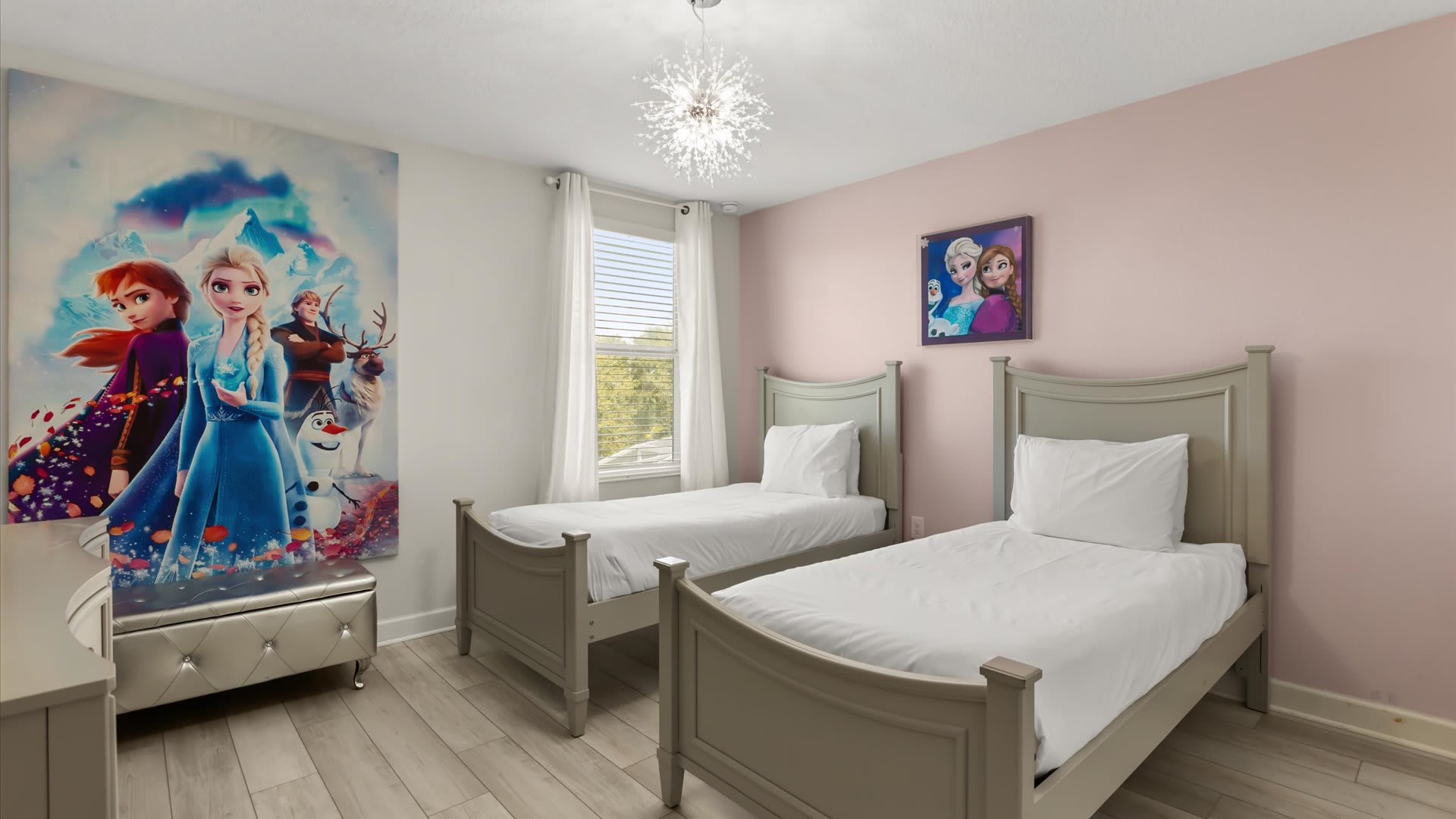 A charming Frozen-themed bedroom features two twin beds with light gray frames and white bedding, set against a light pink wall with a large Frozen mural. A modern sputnik-style chandelier hangs from the ceiling.