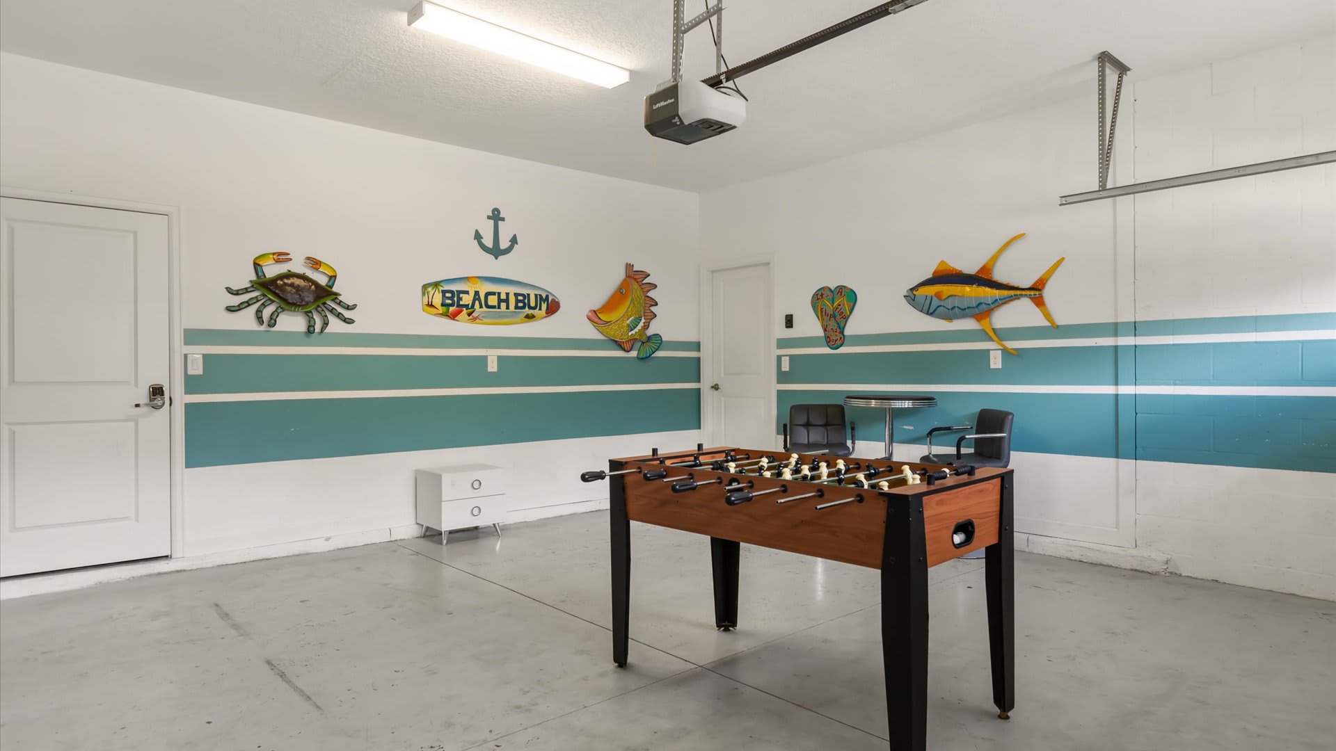 A garage converted into a game room features a foosball table in the center, with white walls decorated with colorful marine-themed metal art and a blue stripe. Two chairs and a small table are in the background.