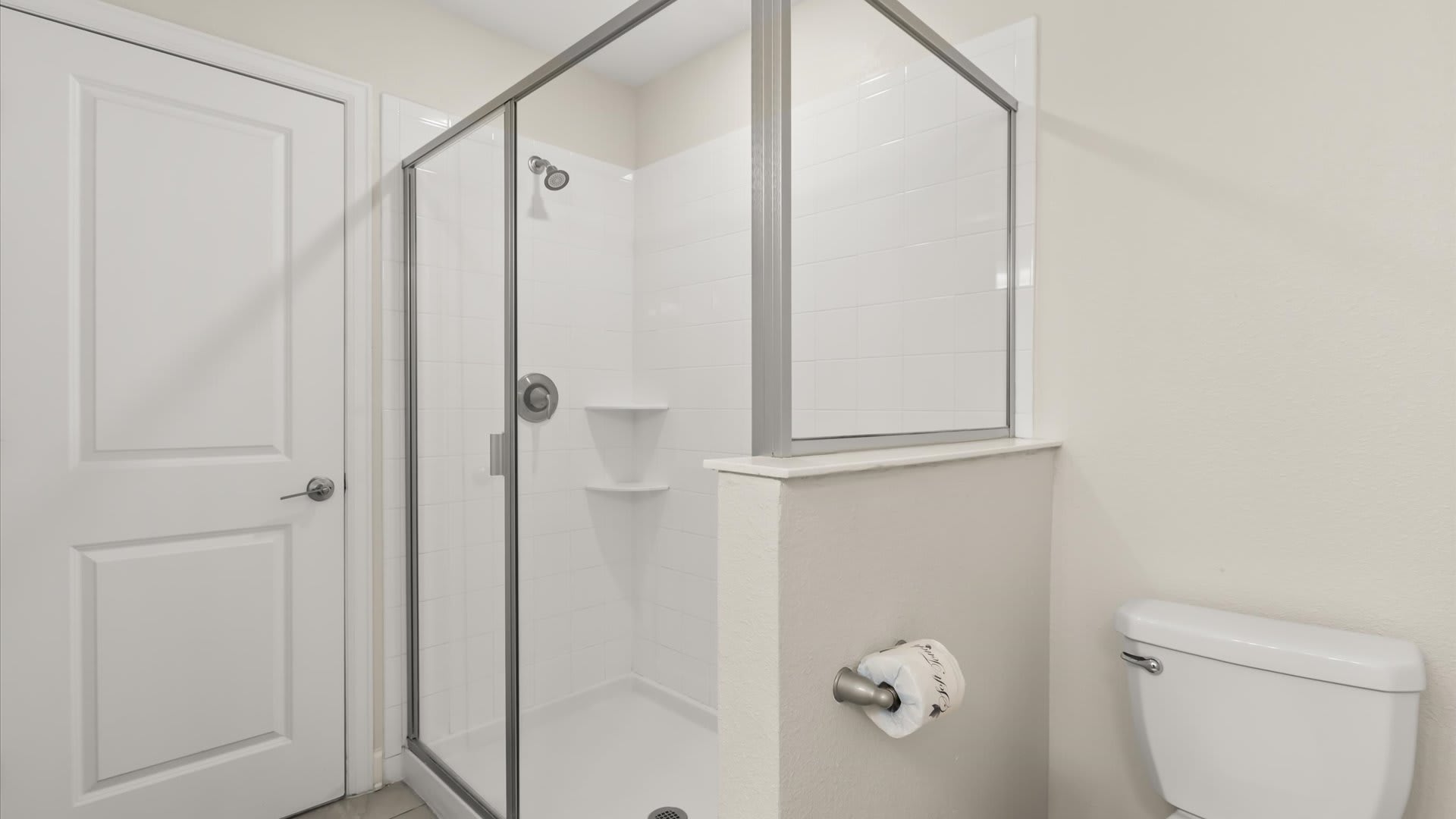 A modern walk-in shower with clear glass panels and white subway tiles is positioned next to a white toilet, separated by a low wall. A roll of toilet paper is mounted on the wall beside the toilet.