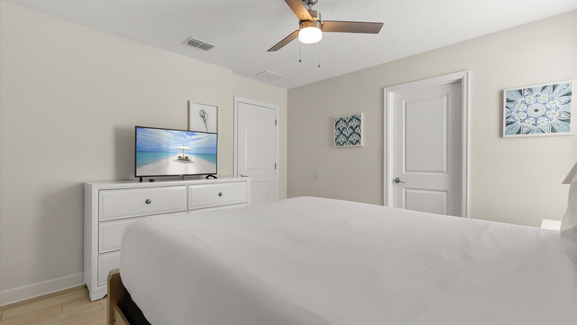 Another view of the bedroom, featuring a white dresser with a flat-screen TV displaying a tranquil beach scene. The room has light wood-look flooring and light-colored walls adorned with decorative artwork.
