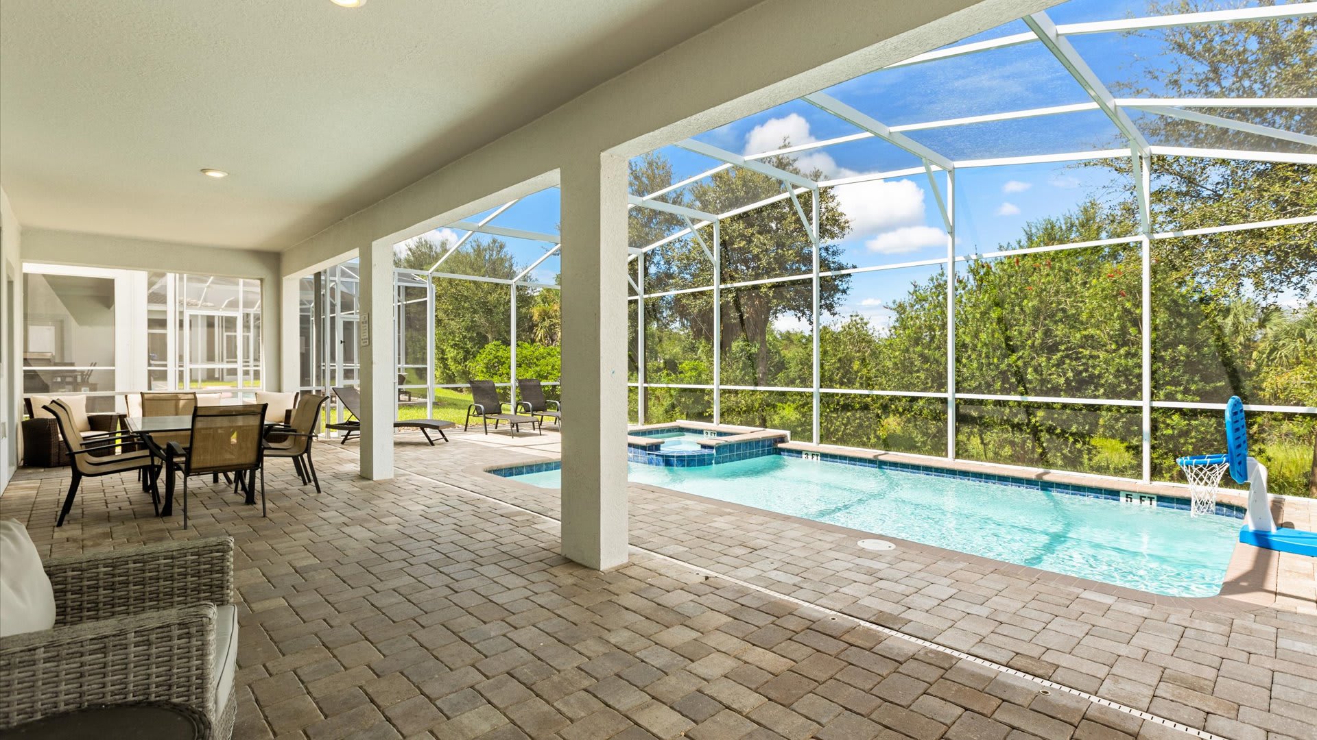 A wide-angle view of the screened-in patio shows a covered area with outdoor seating and a dining table, overlooking a swimming pool and hot tub. Beyond the screen, dense green foliage provides privacy.