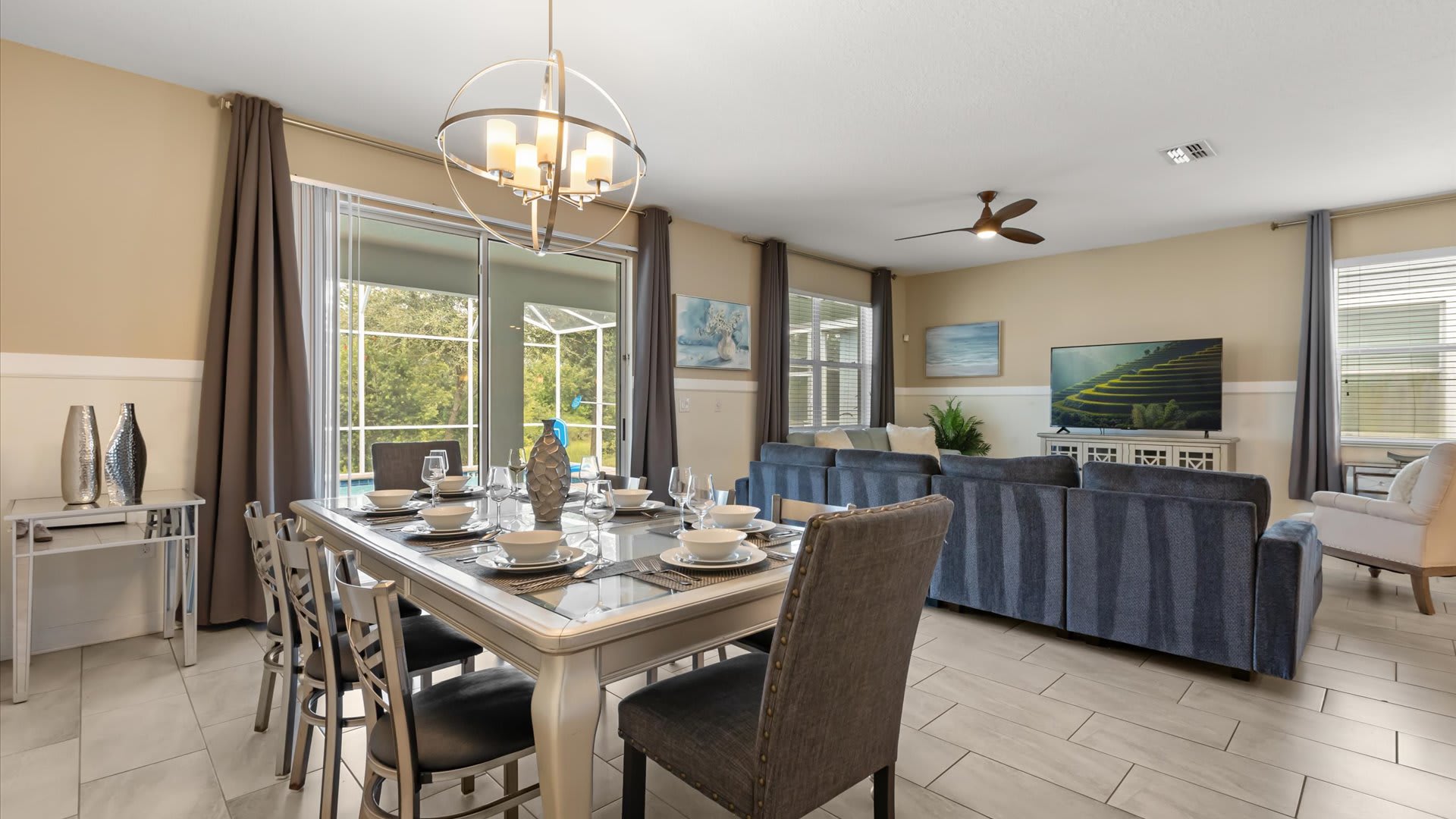 Open-concept space combining a dining area with a modern chandelier and a living room with a large sectional sofa, both featuring light tile flooring and access to an outdoor pool through sliding doors.