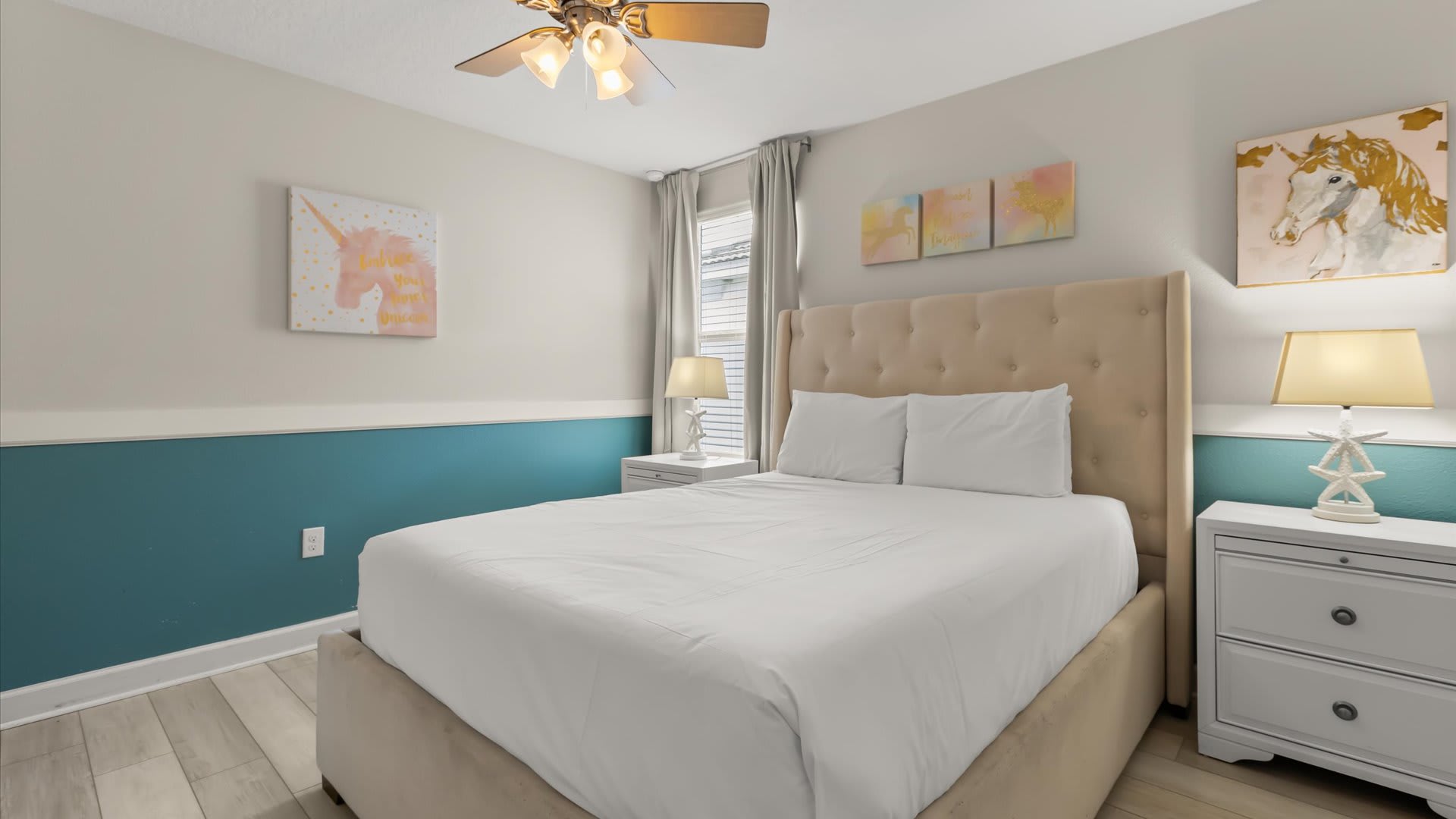 Vibrant bedroom with a beige upholstered queen bed, white bedding, and two white nightstands with lamps. A distinctive teal wainscoting accent wall adds a pop of color, complemented by unicorn and abstract art.