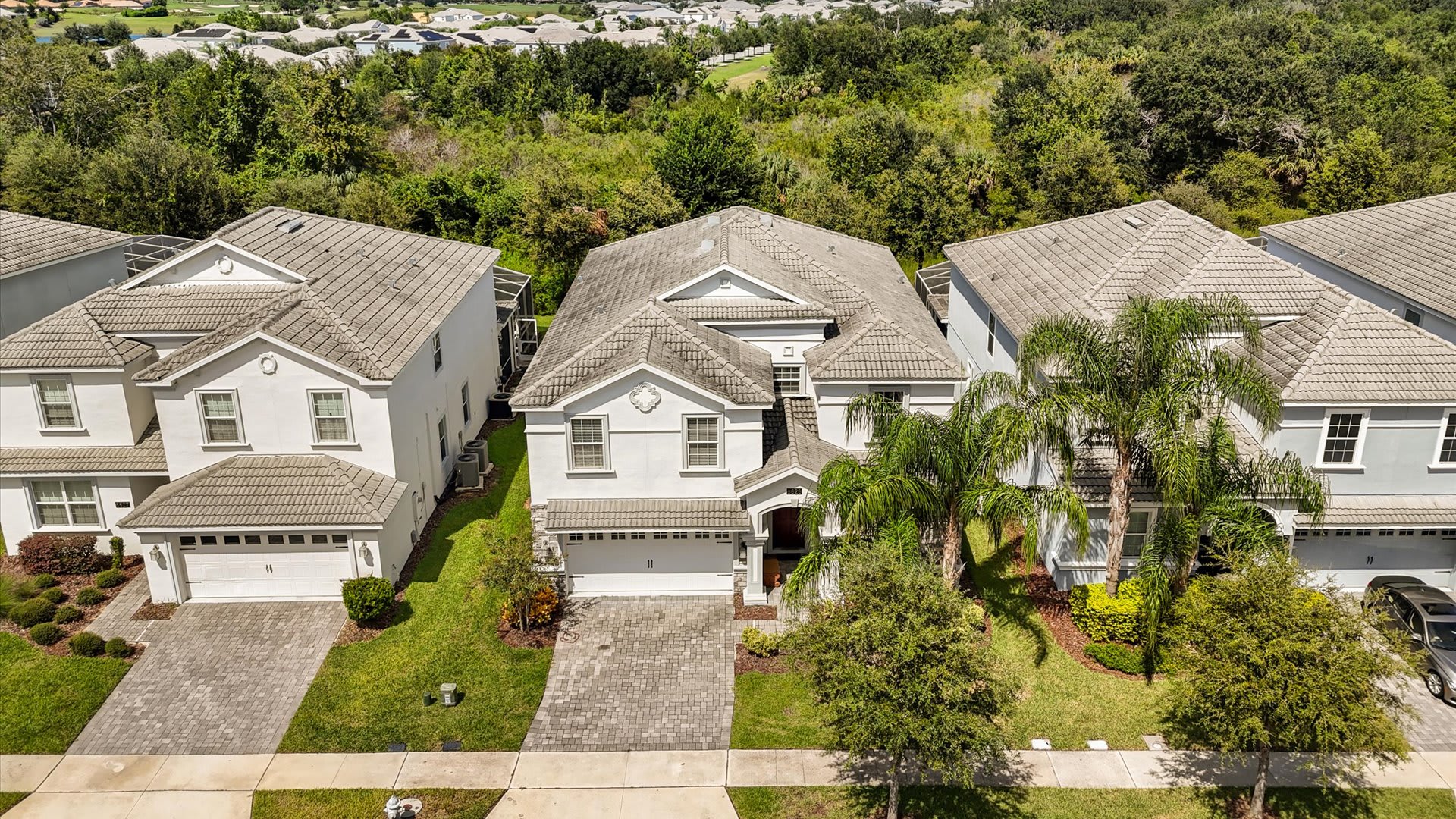 Overhead aerial view of a large, light-colored two-story house with a gray tiled roof, a paved driveway, and green landscaping, situated within a residential neighborhood.